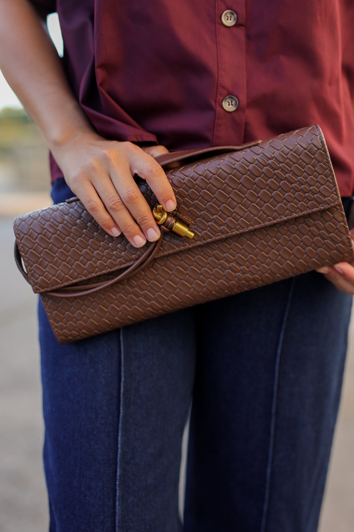 A person holding a brown woven leather clutch bag with a gold clasp, wearing a burgundy button-up shirt and blue jeans.