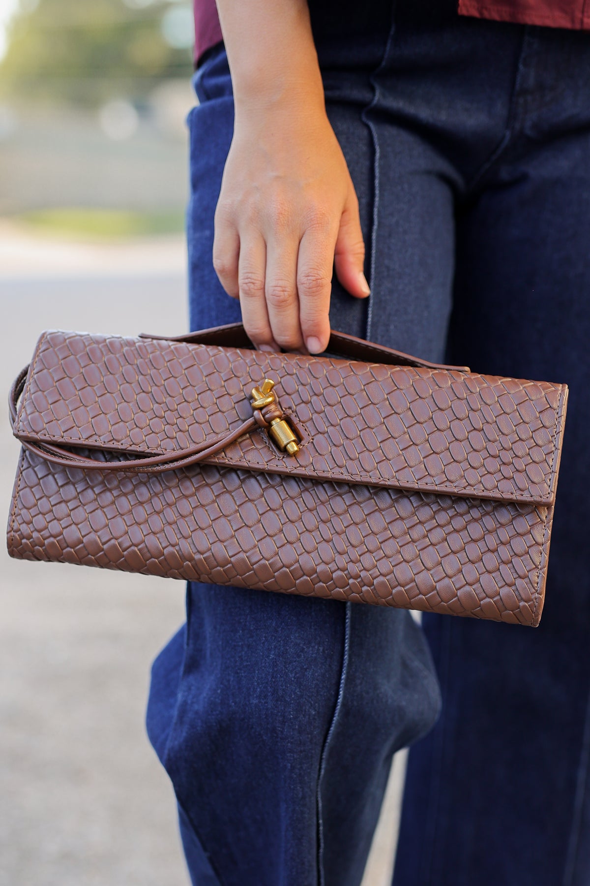 A brown textured clutch purse with a gold-toned toggle clasp, held by a person wearing blue jeans.