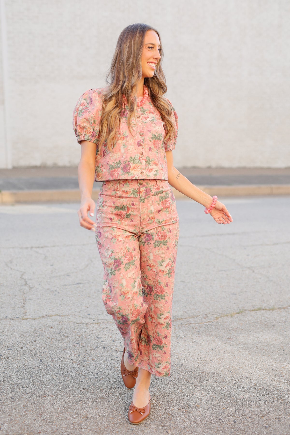 A woman walking outdoors wearing a matching dusty pink floral two-piece set with puff sleeves and cuffed pants, paired with brown flat shoes.