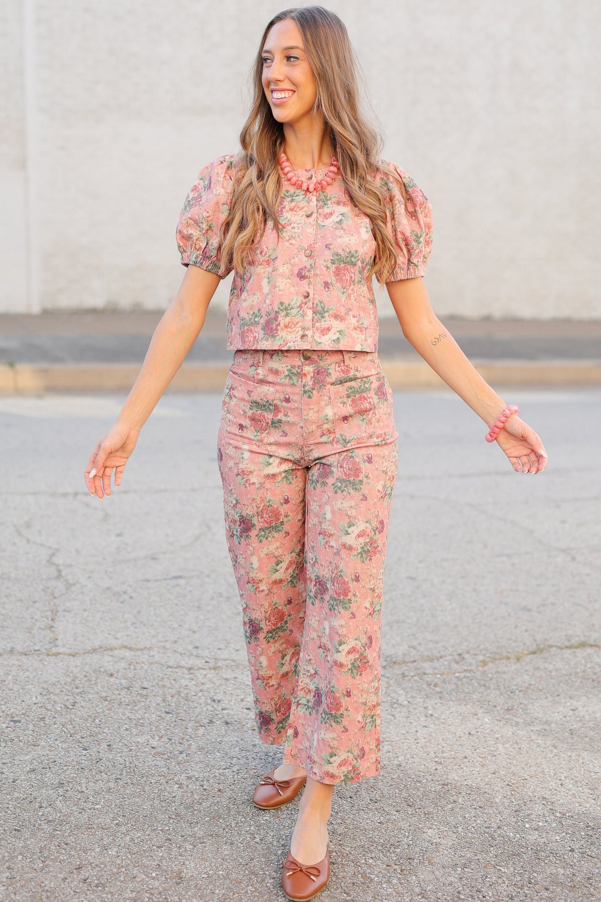 A woman wearing a matching two-piece floral set with puff sleeves and cropped pants, walking outdoors on a paved surface against a light-colored wall.
