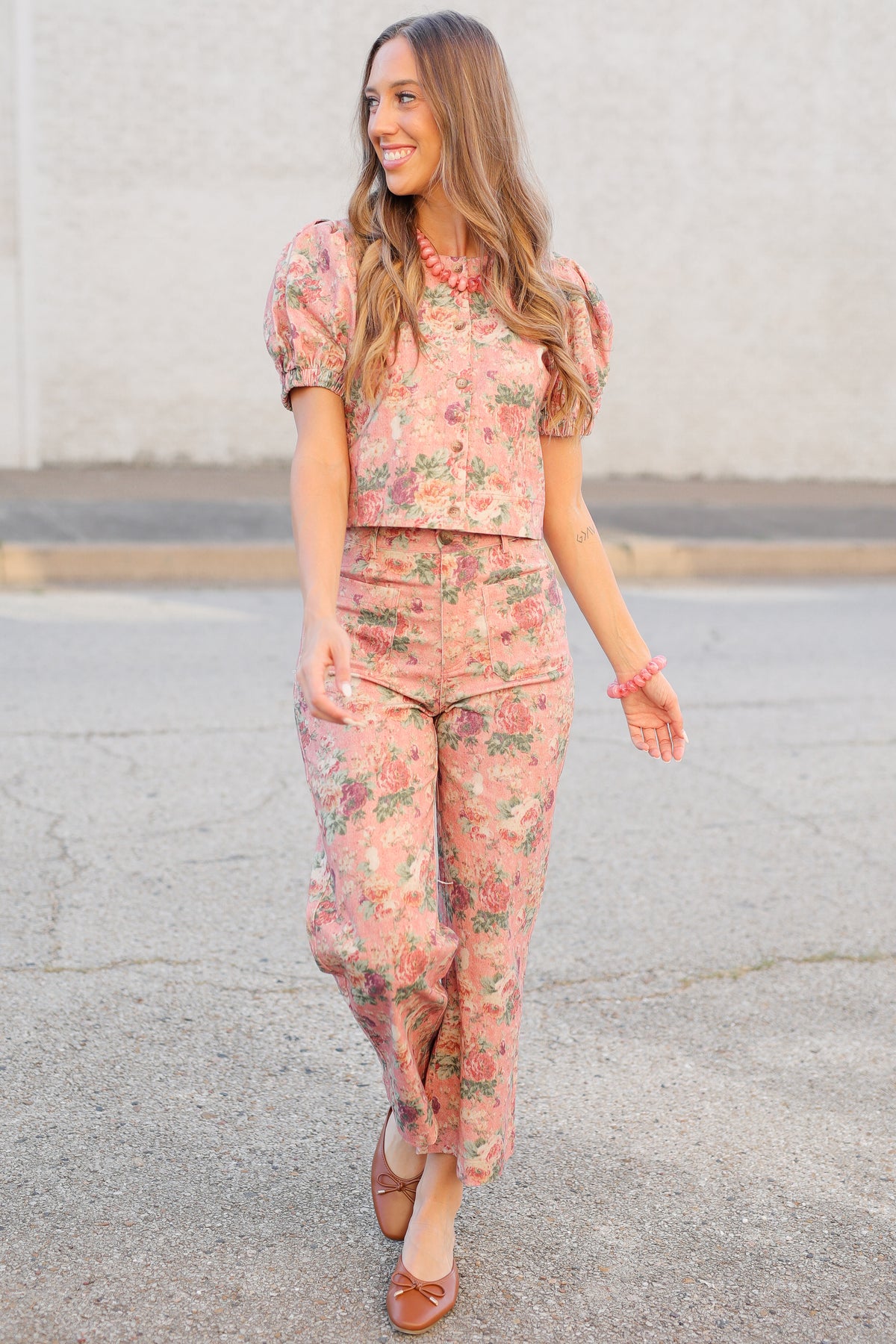 A woman wearing a matching pink floral puff-sleeve top and high-waisted wide-leg pants set, walking outdoors on a paved surface against a light concrete wall.