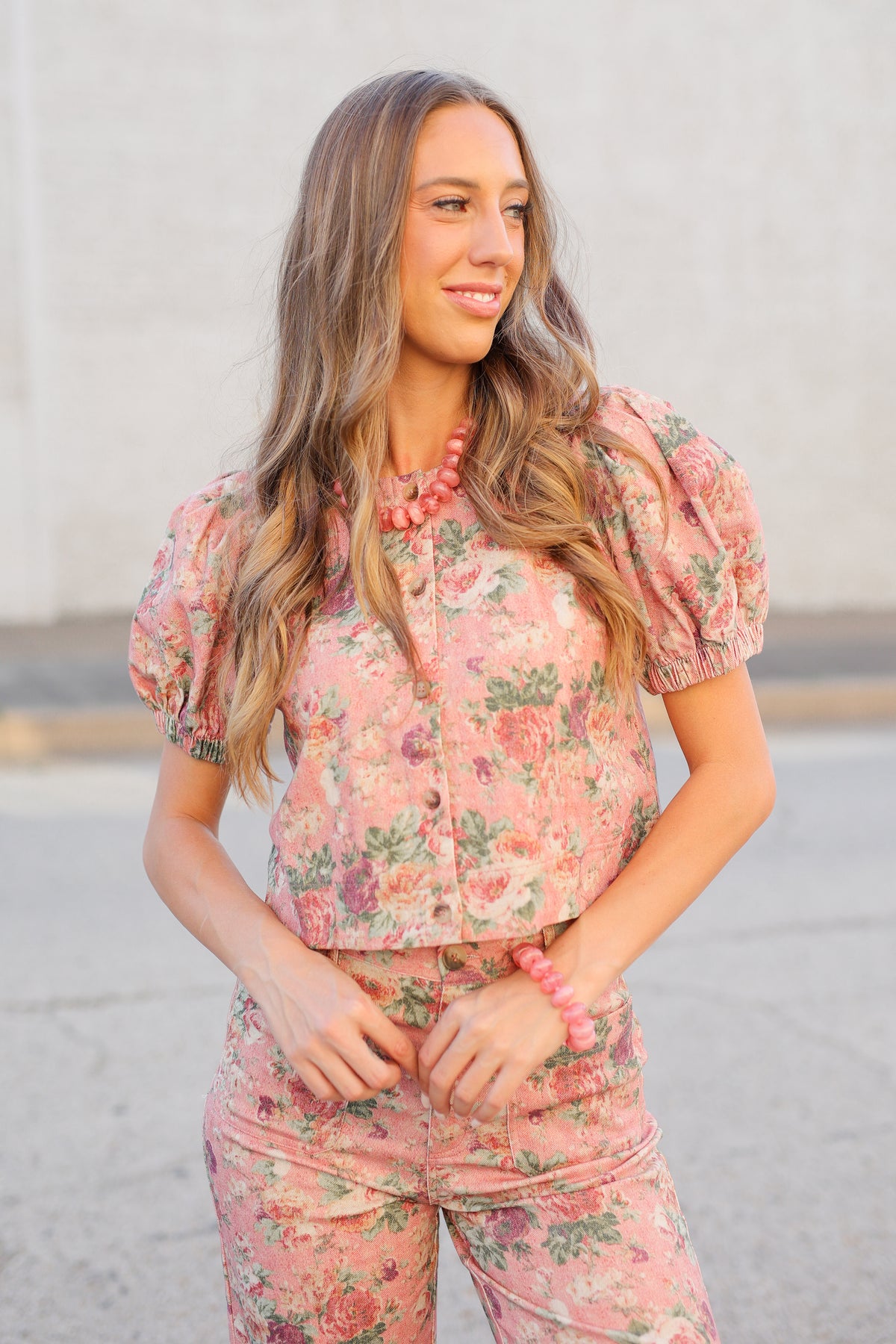 A woman wearing a pink floral puff-sleeve button-up shirt and matching floral pants, standing outdoors against a light neutral wall.