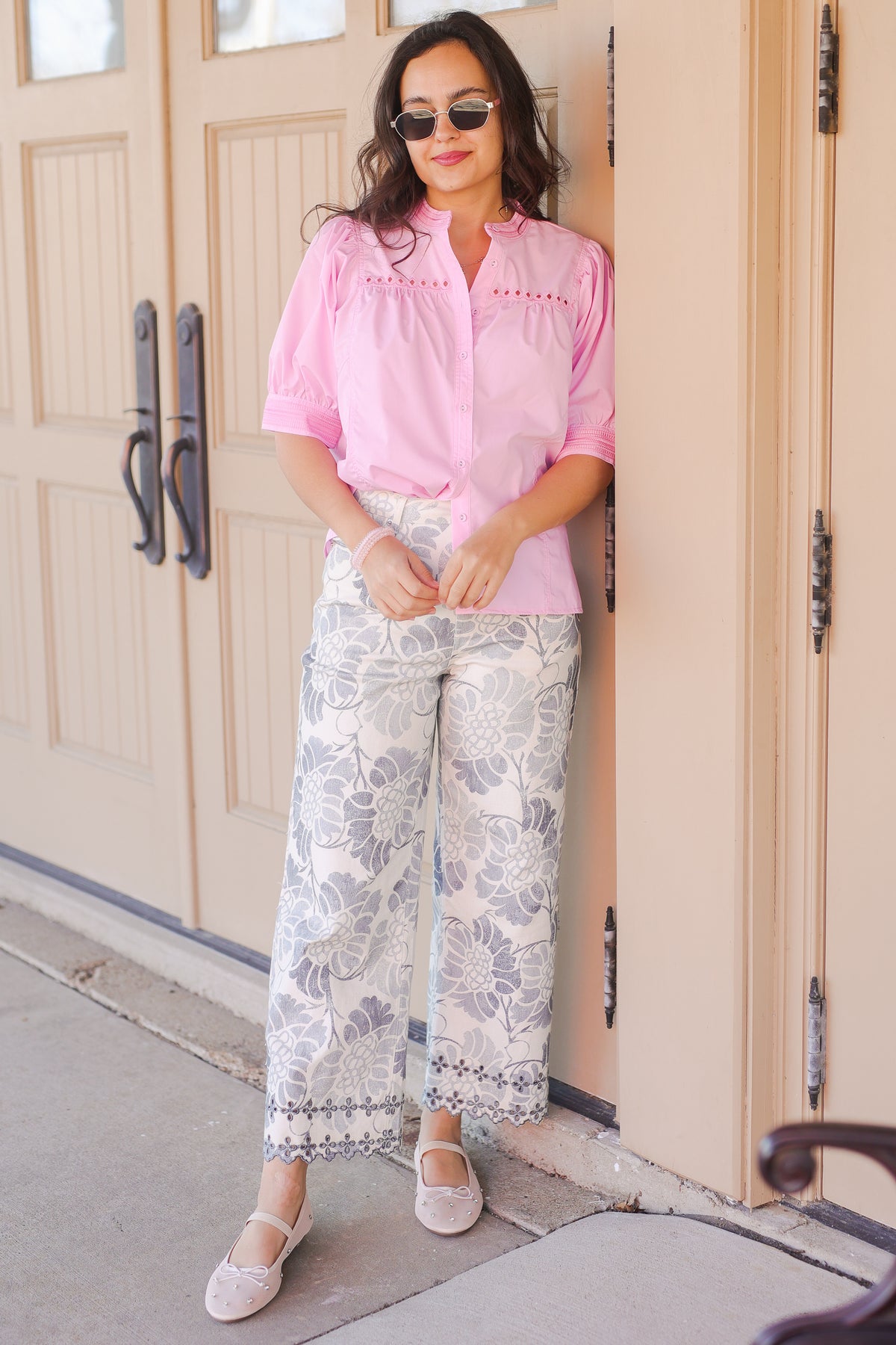 Woman wearing a pink blouse and floral pants standing in front of a door.