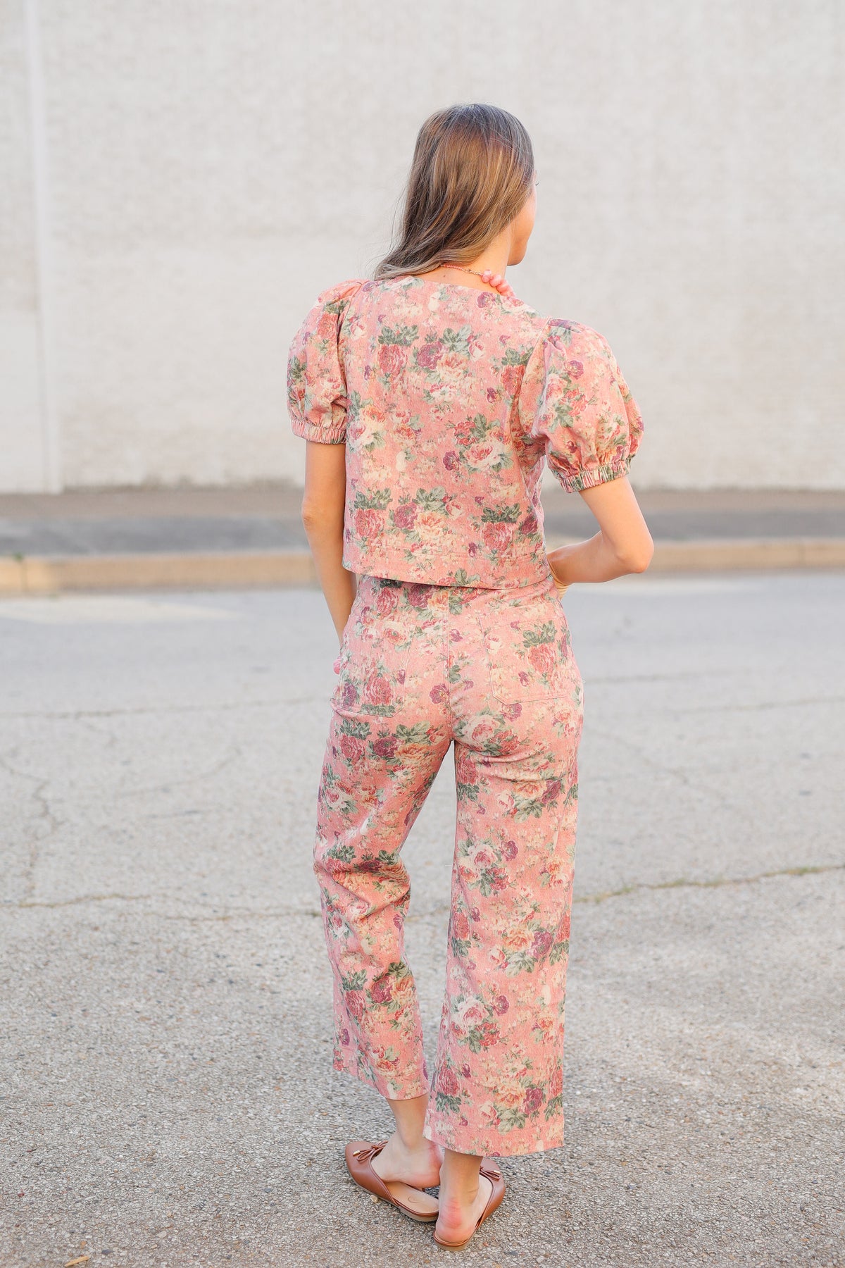 A woman viewed from behind wearing a matching pink floral puff-sleeve top and cropped pants set, standing on a paved surface against a light concrete wall.