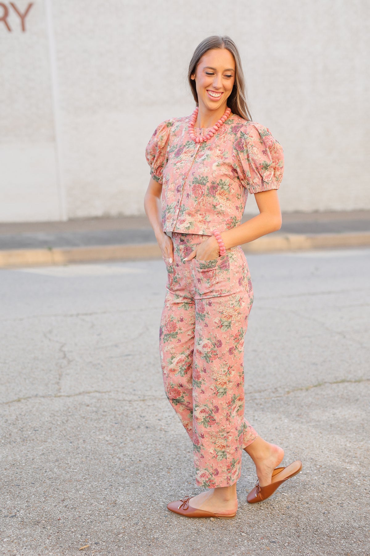 A woman wearing a pink floral puff-sleeve top and matching wide-leg pants, standing outdoors on a paved surface against a light-colored wall.