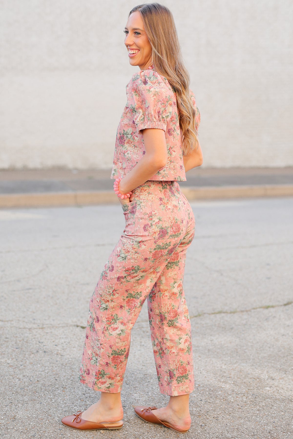 A woman wearing a matching two-piece floral lounge set in pink, featuring a short-sleeve crop top and cropped pants, standing outdoors on a paved surface.