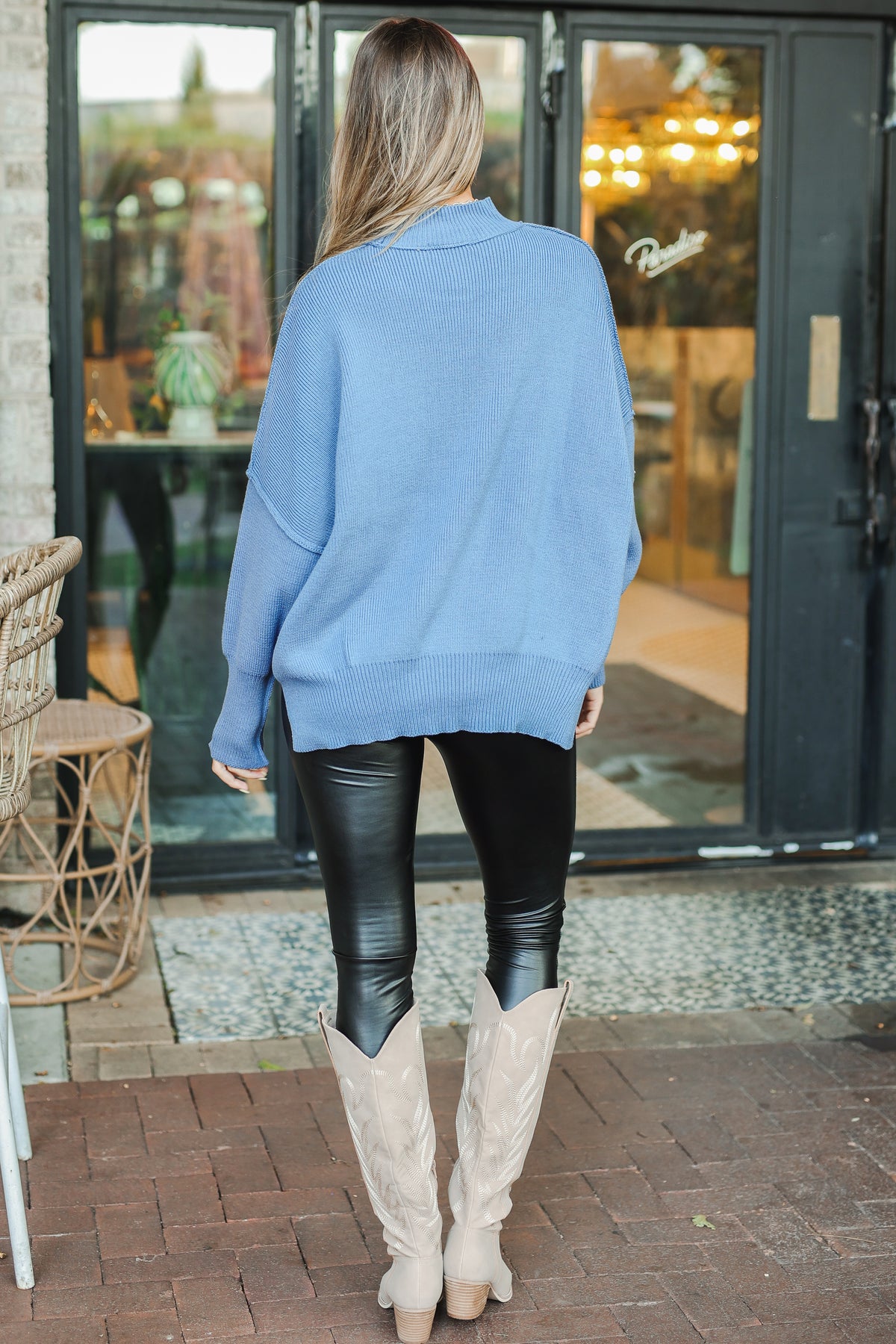 Person wearing a light blue sweater, black leggings, and beige boots standing in front of a glass door.