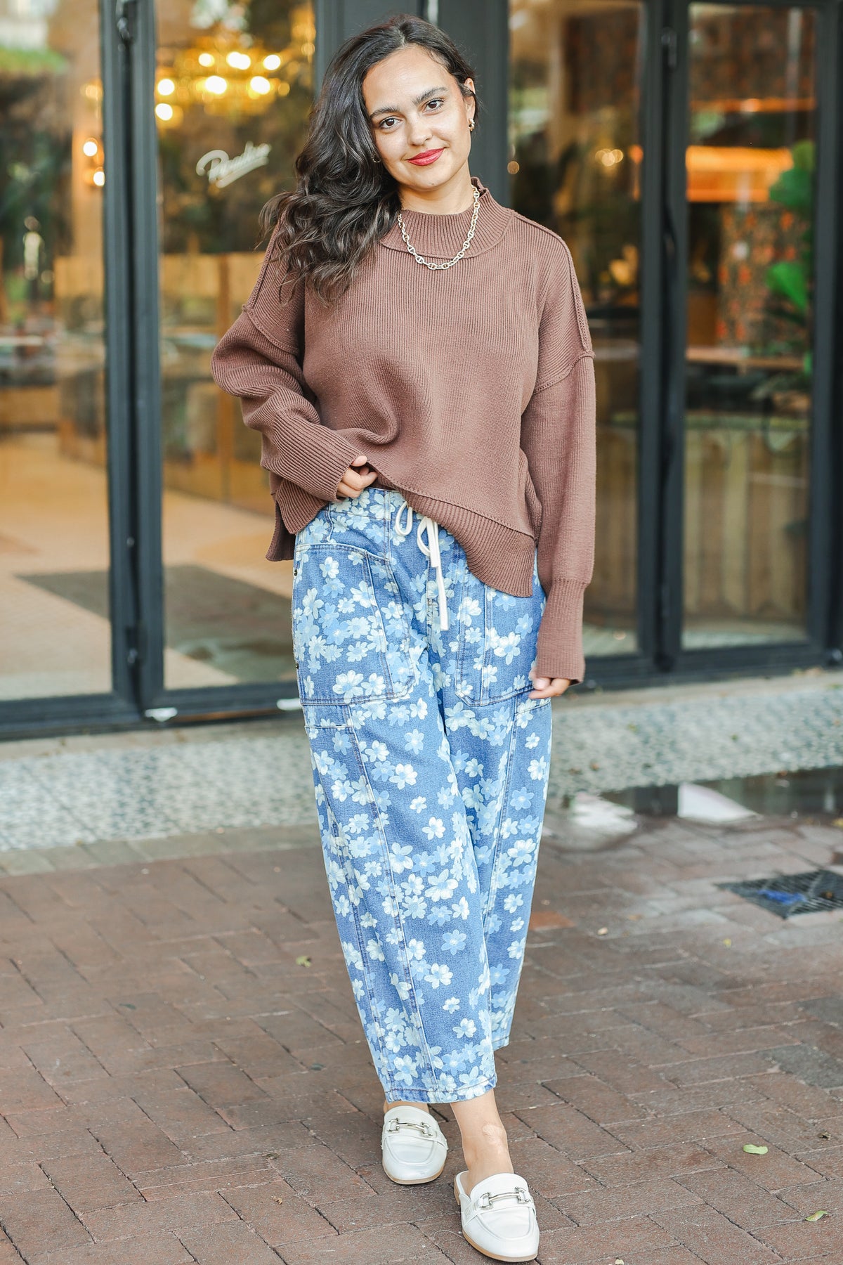 Woman wearing a brown sweater and blue floral pants standing on a sidewalk.