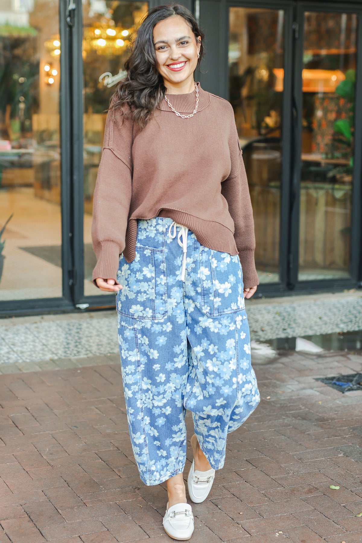 Woman wearing a brown sweater and blue floral pants standing outdoors.