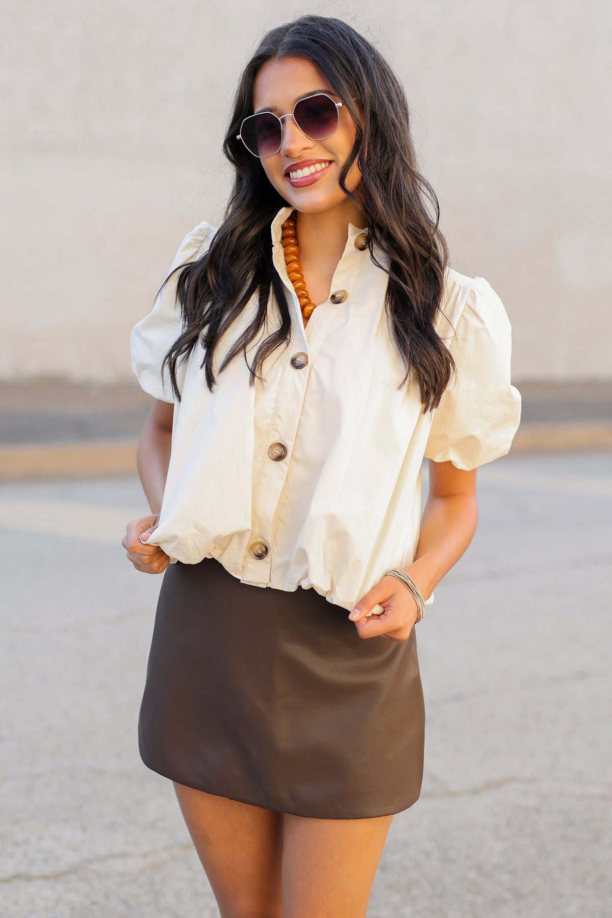 A woman wearing a cream puff-sleeve button-front blouse and a brown mini skirt, standing outdoors against a neutral wall.