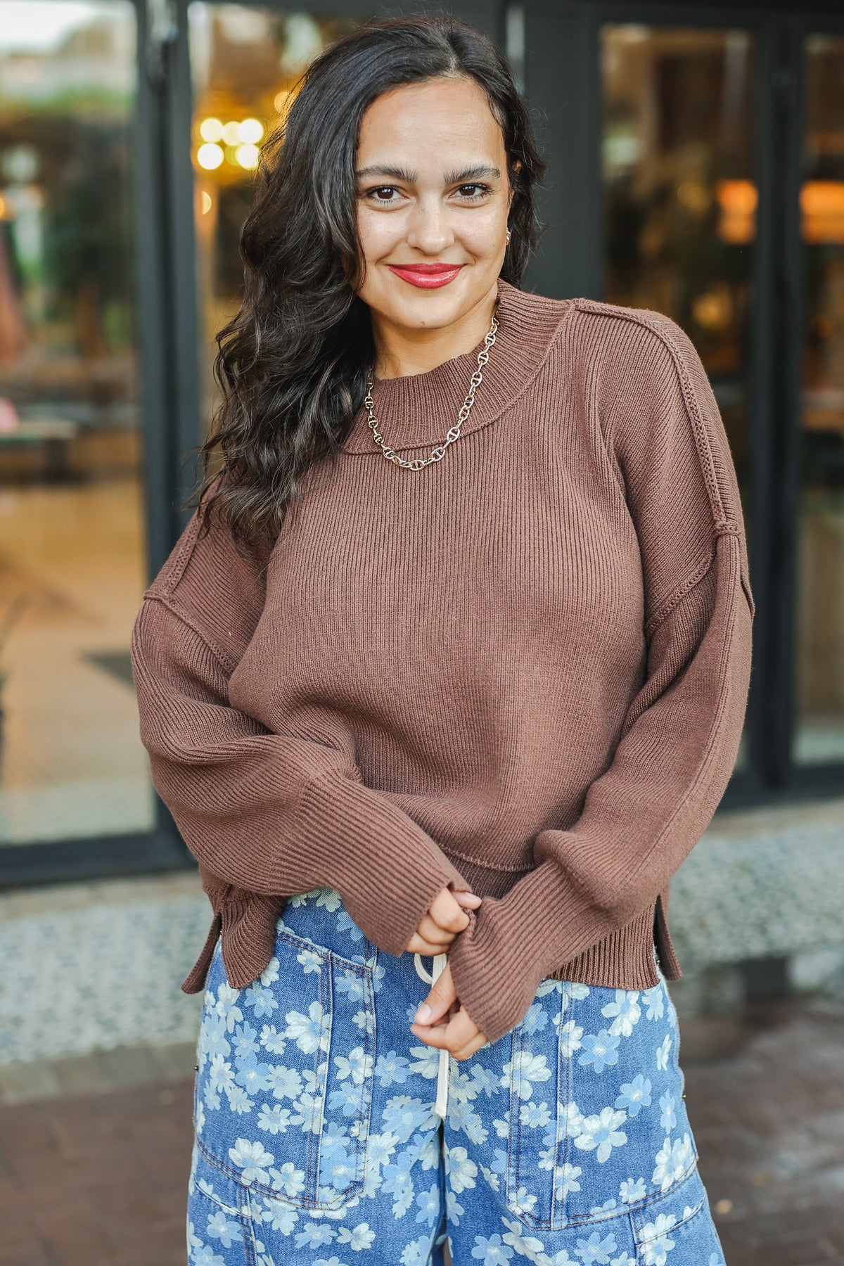 Woman wearing a brown sweater and blue floral skirt standing outdoors.