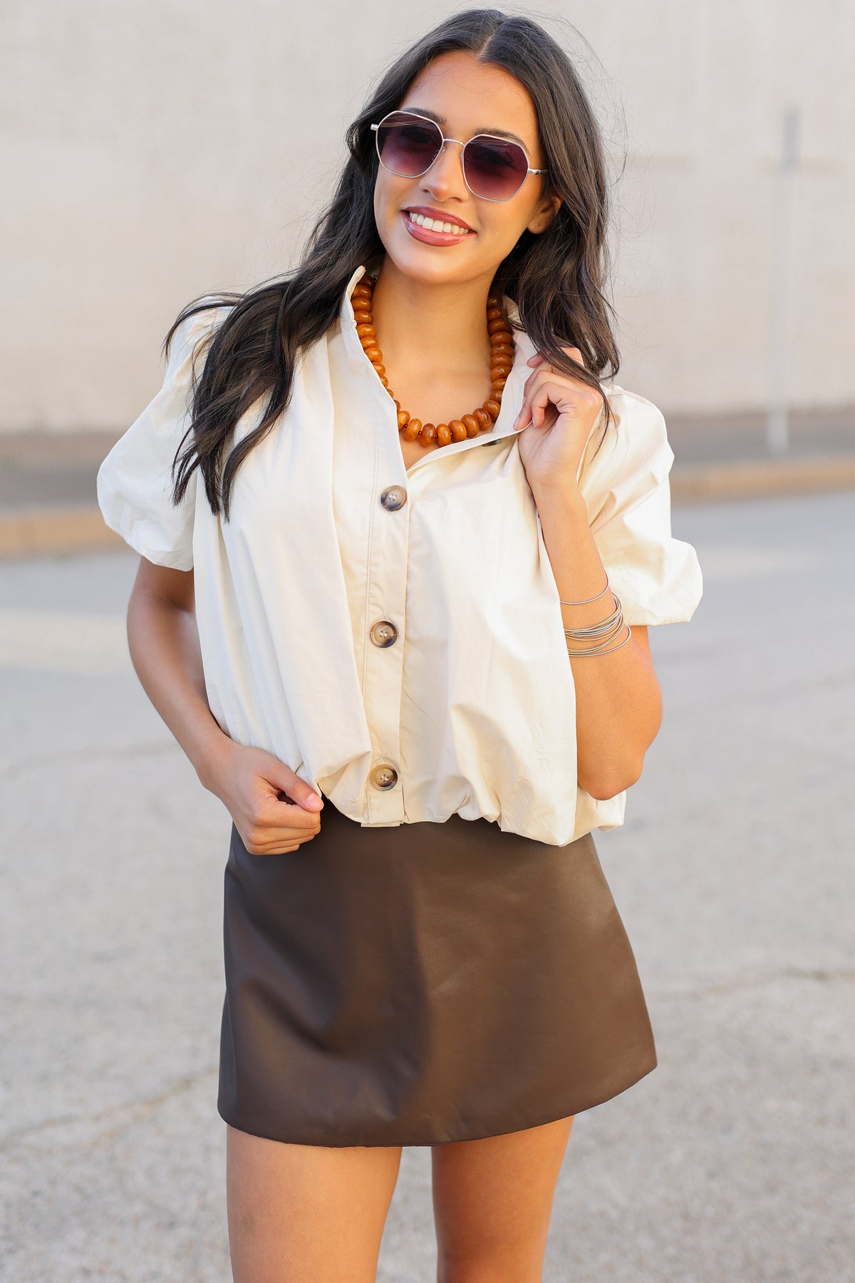 A woman wearing a cream puff-sleeve button-up shirt and a brown mini skirt, accessorized with sunglasses, a chunky orange beaded necklace, and bracelets, standing outdoors against a neutral concrete wall.