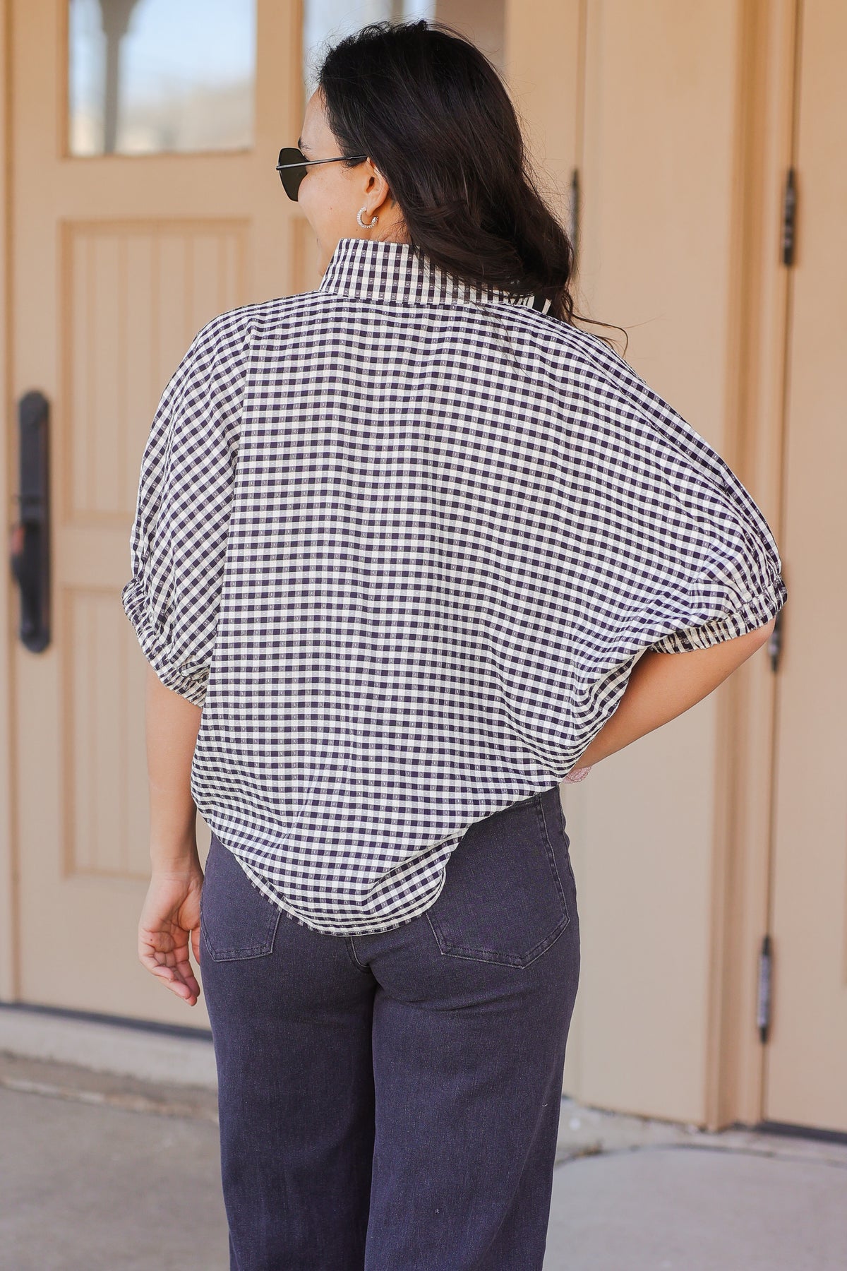Woman wearing a black and white checkered blouse and dark pants standing in front of a door.
