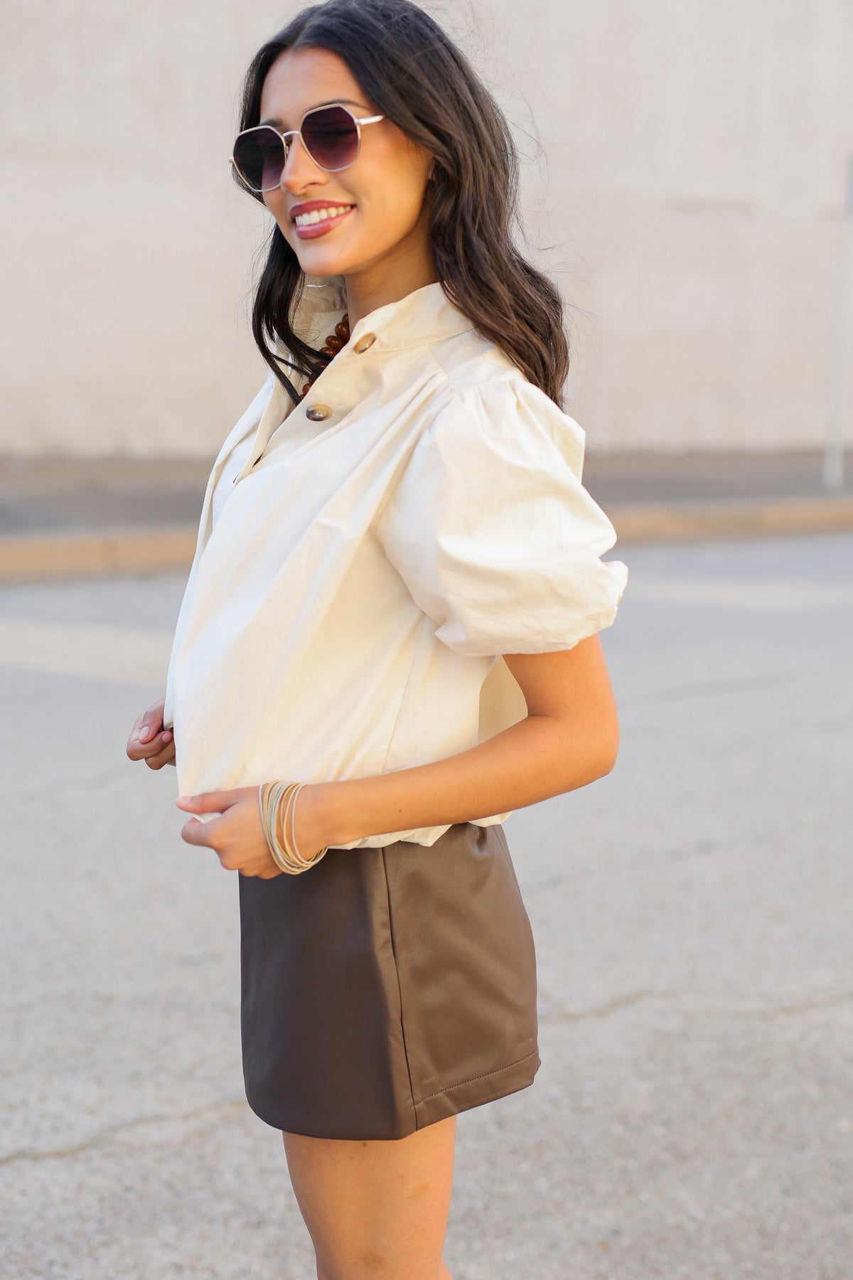 A woman wearing a cream puff-sleeve blouse and a brown faux-leather mini skirt, standing outdoors on a paved surface against a beige wall.