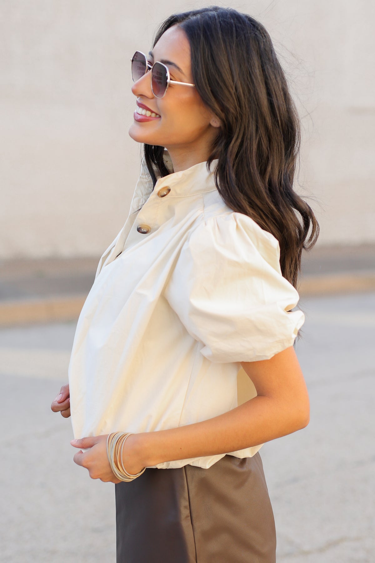 A woman wearing a cream puff-sleeve blouse with a high buttoned collar and brown satin trousers, accessorized with sunglasses and gold bracelets, standing outdoors against a beige wall.