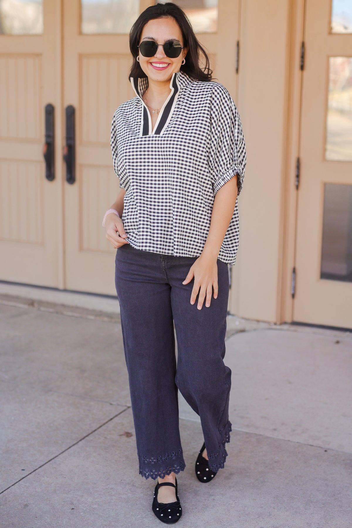 Woman wearing a checkered blouse and dark pants standing in front of a beige building.