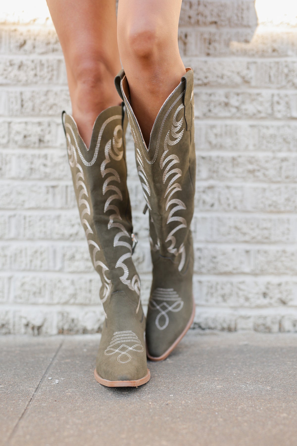 A person wearing knee-high western boots in a muted olive green color with white embroidered detailing, standing on a concrete surface in front of a white brick wall.