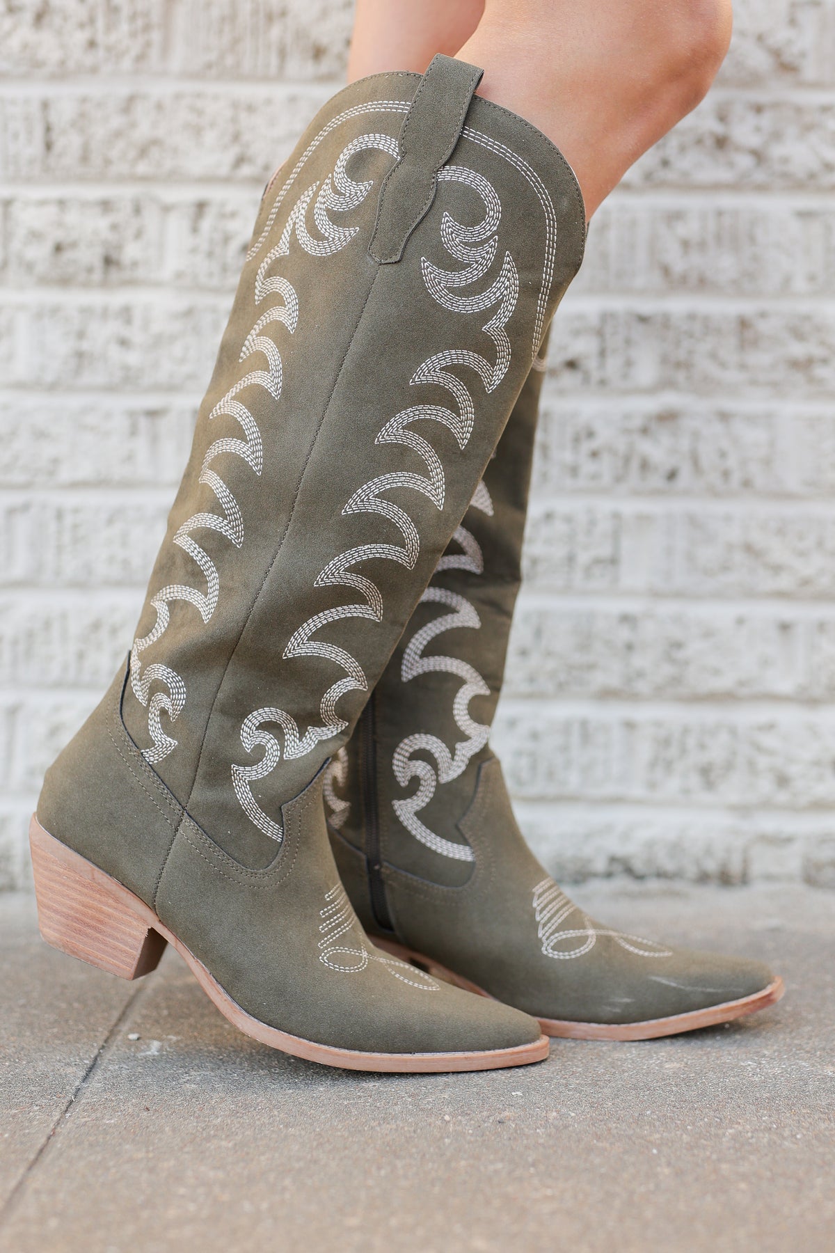 A pair of knee-high western boots in olive green suede with white embroidered scrollwork, shown on a model standing against a light gray brick wall.