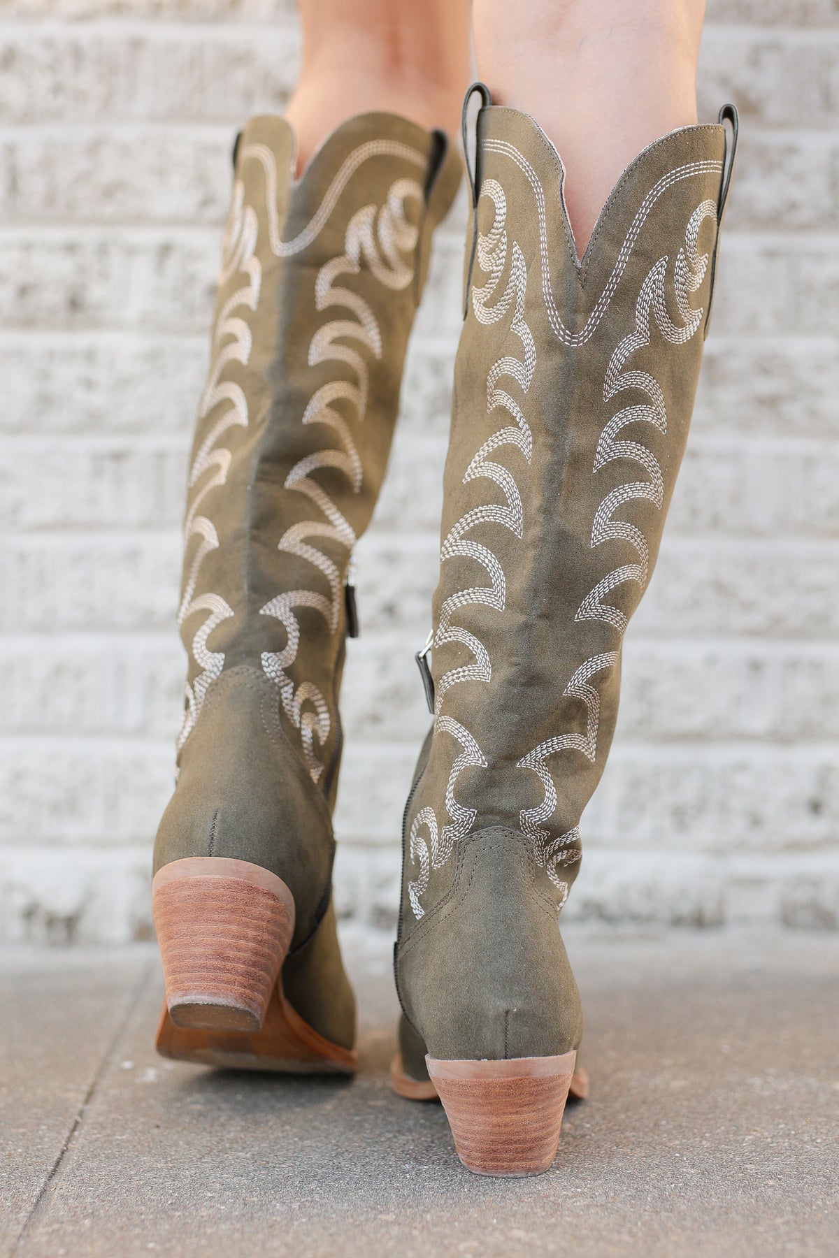 A pair of knee-high western boots in taupe suede with white embroidered stitching, shown on a model's feet against a white brick wall.