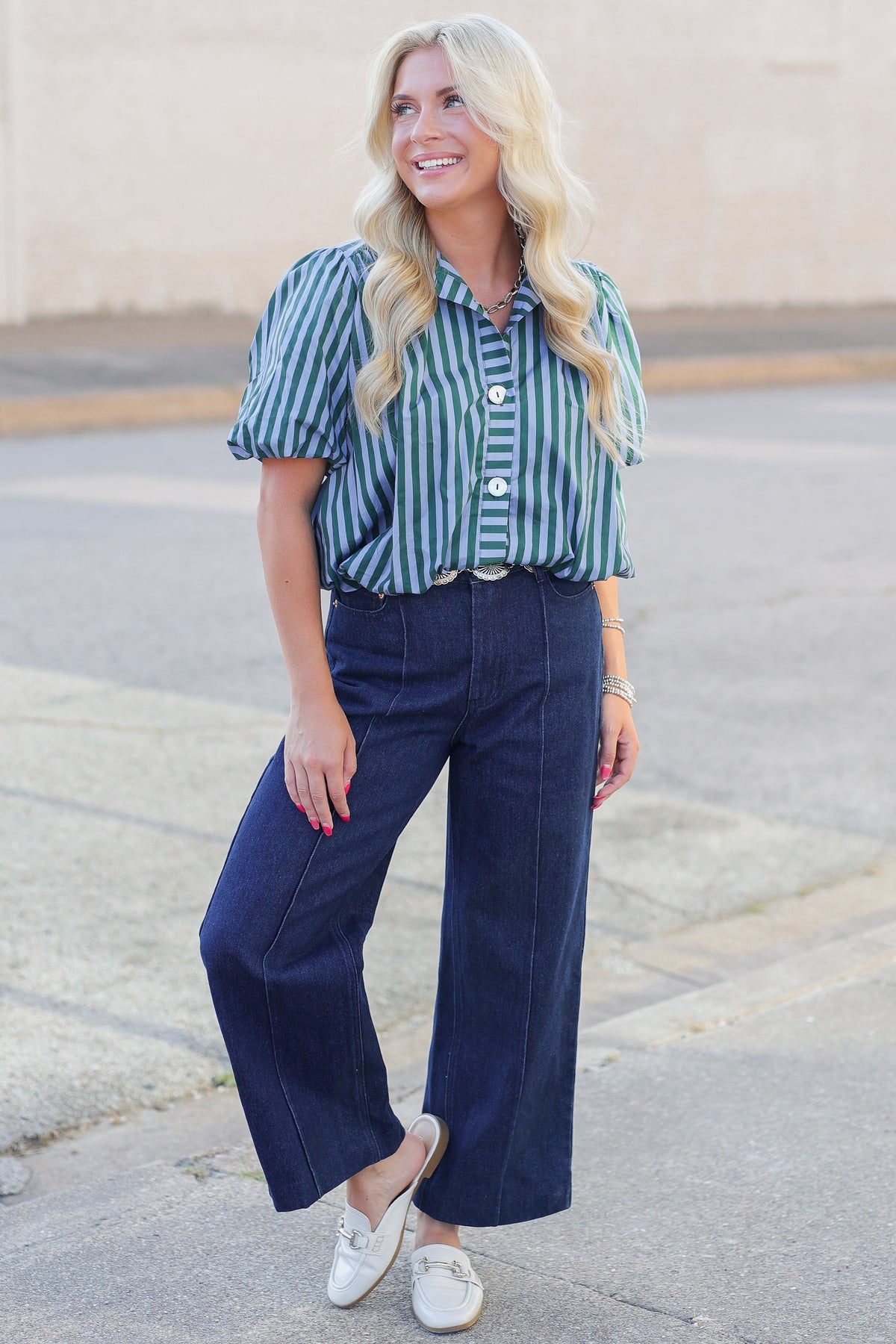 A woman stands outdoors wearing a green and white vertically striped short-sleeve button-up shirt, dark blue wide-leg jeans, and white slip-on shoes.