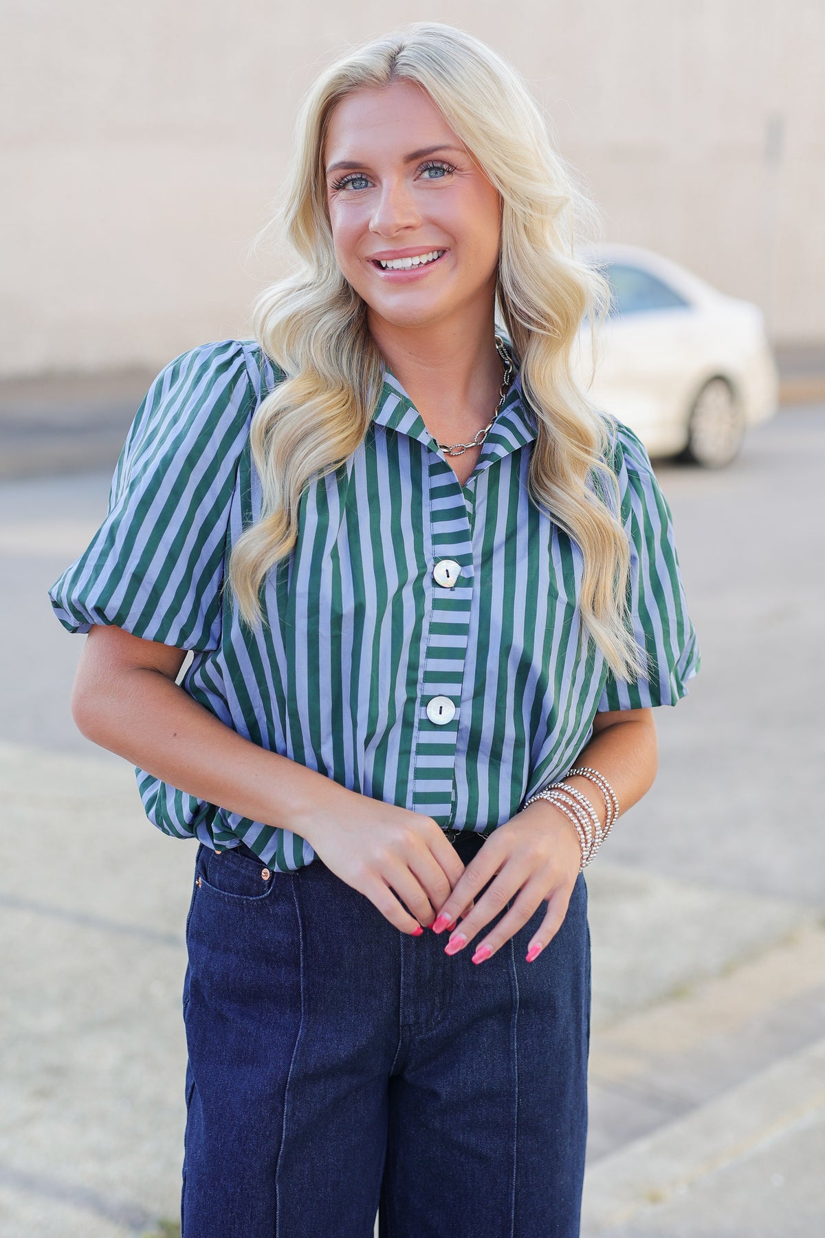 A smiling blonde woman wearing a green and white vertically striped short-sleeve button-up shirt with dark blue jeans, standing outdoors in front of a neutral-toned wall and a blurred white car.