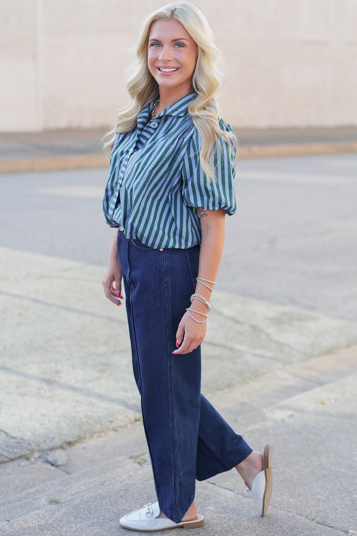 A woman wearing a green and white vertically striped short-sleeve blouse with puff sleeves, paired with dark blue wide-leg jeans and white loafers, standing outdoors on a paved walkway.