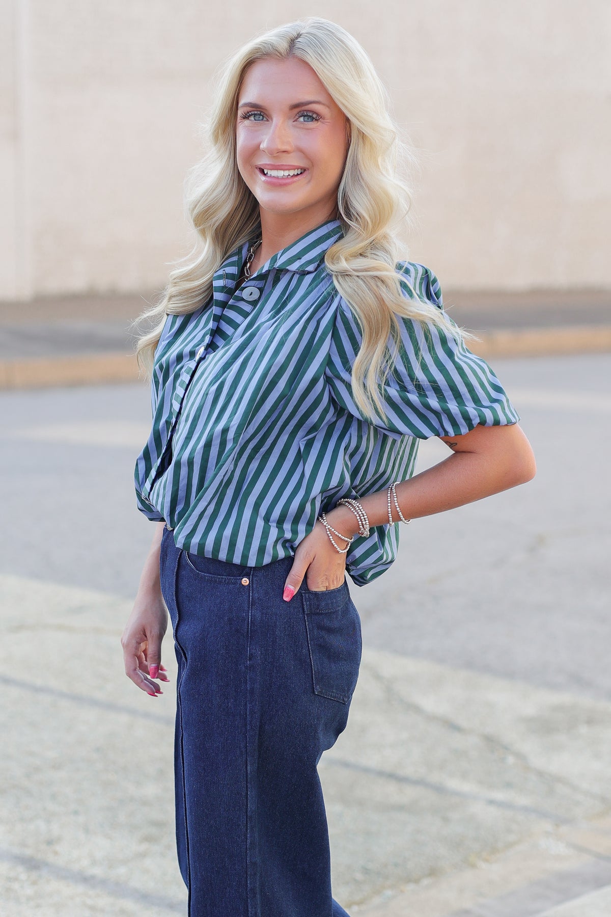 A smiling blonde woman wearing a green and navy blue vertically striped short-sleeve blouse with puff sleeves, paired with dark blue jeans, standing outdoors against a beige wall.