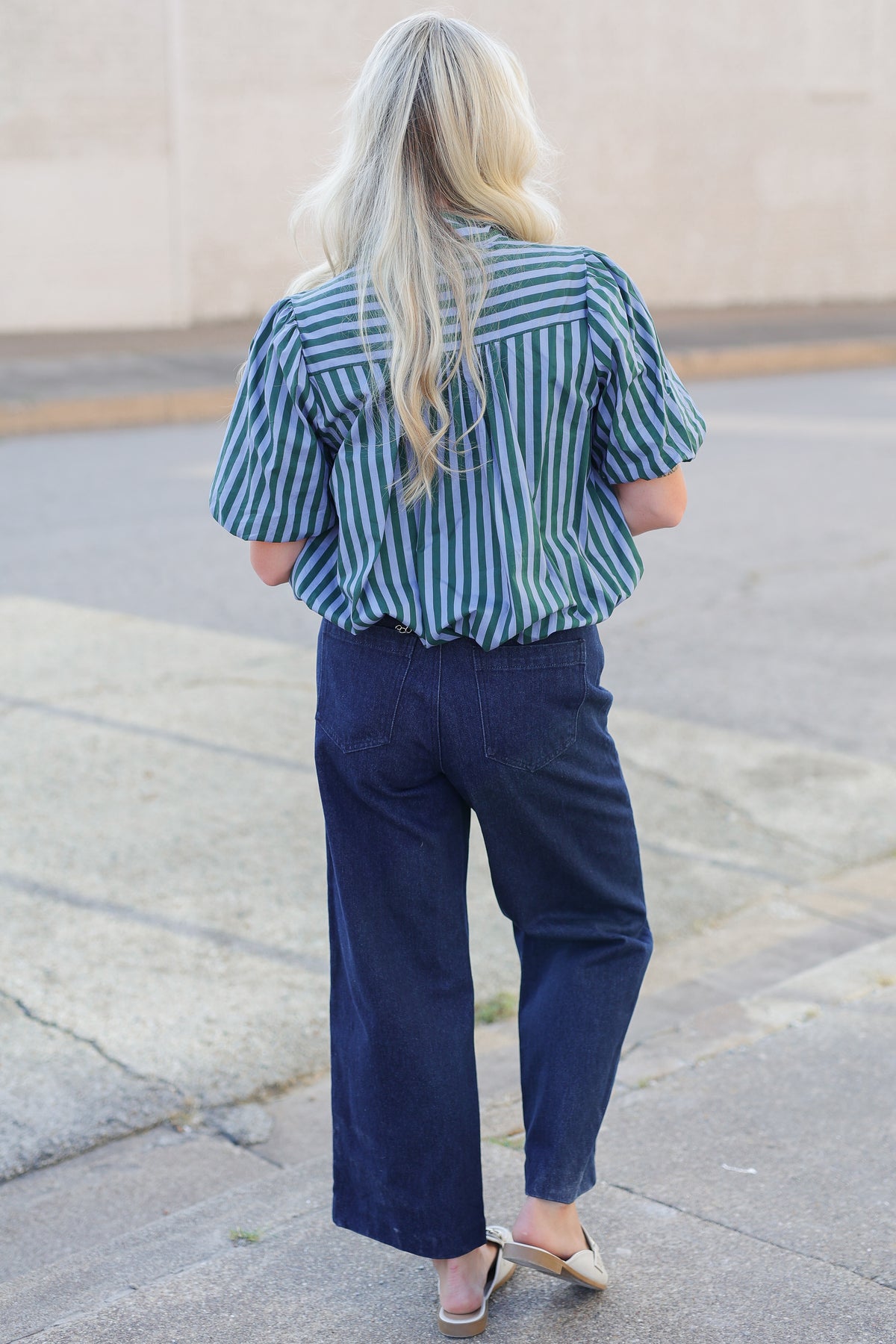 A woman viewed from behind wearing a blue and green vertically striped short-sleeve button-up shirt and dark navy wide-leg pants, standing on a paved outdoor walkway.