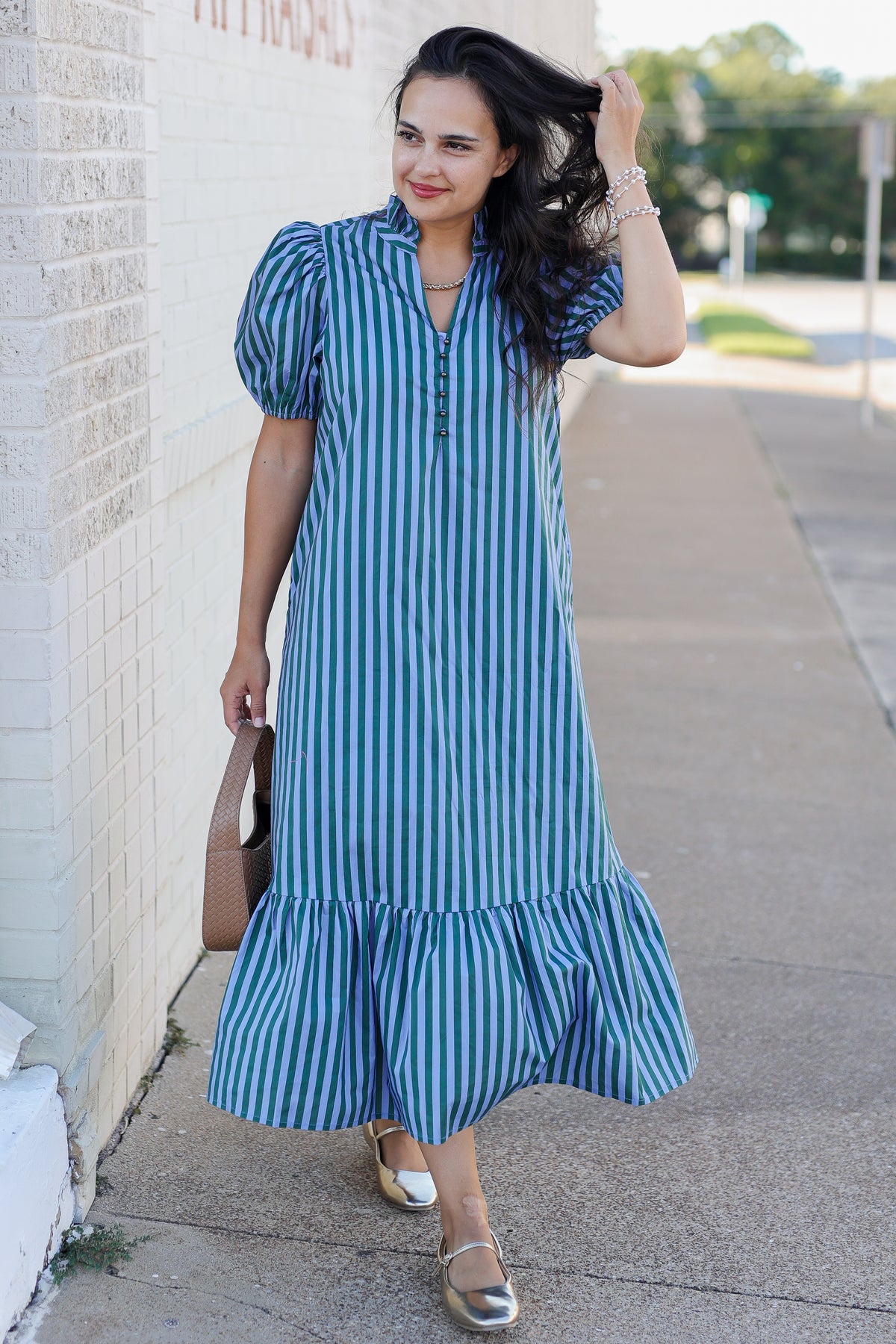 A woman wearing a blue and white vertically striped midi dress with puff sleeves and a ruffled hem, walking on a sidewalk next to a light-colored brick wall.