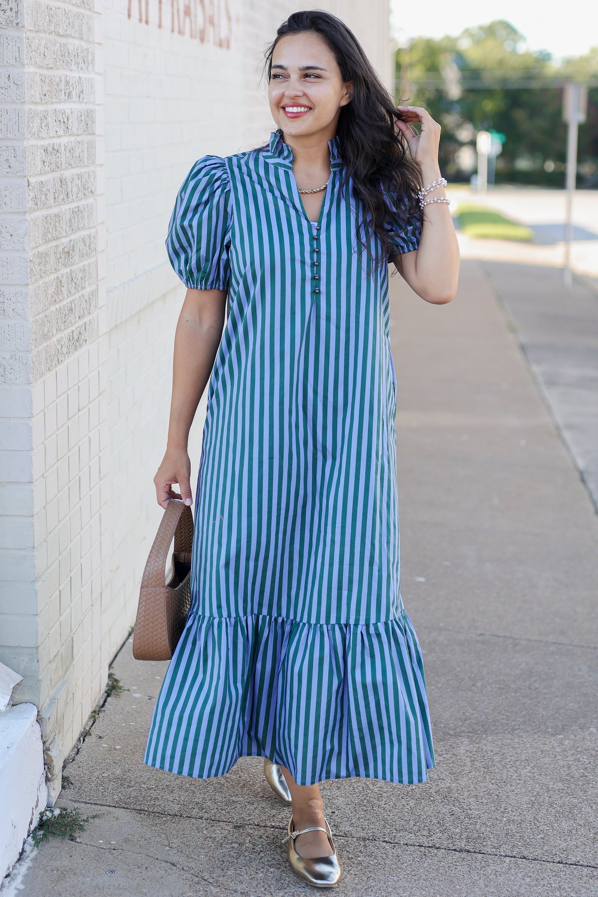 A woman wearing a blue and white vertically striped midi dress with puff sleeves and a ruffled hem, walking on a sidewalk next to a light brick wall.
