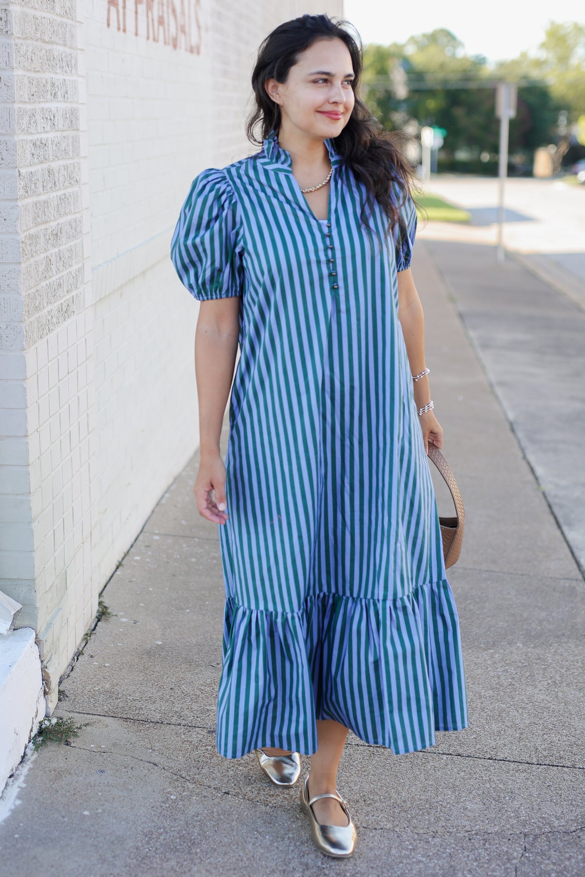 A woman wearing a blue and white vertically striped midi dress with puff sleeves and a tiered hem, walking on a sidewalk next to a white brick wall.