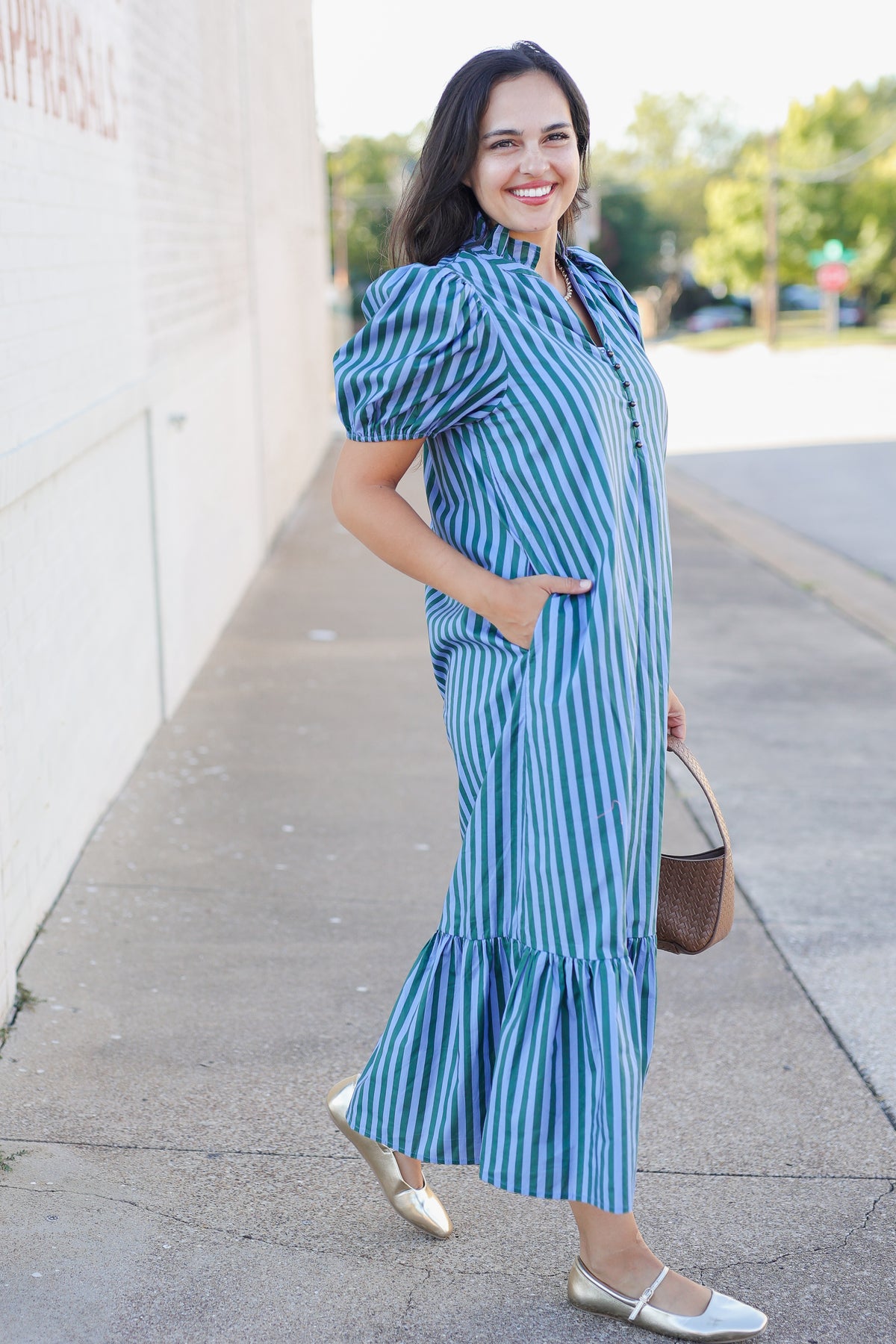 A woman wearing a blue and white vertically striped midi dress with puff sleeves and a ruffled hem, standing on a sidewalk next to a white building.