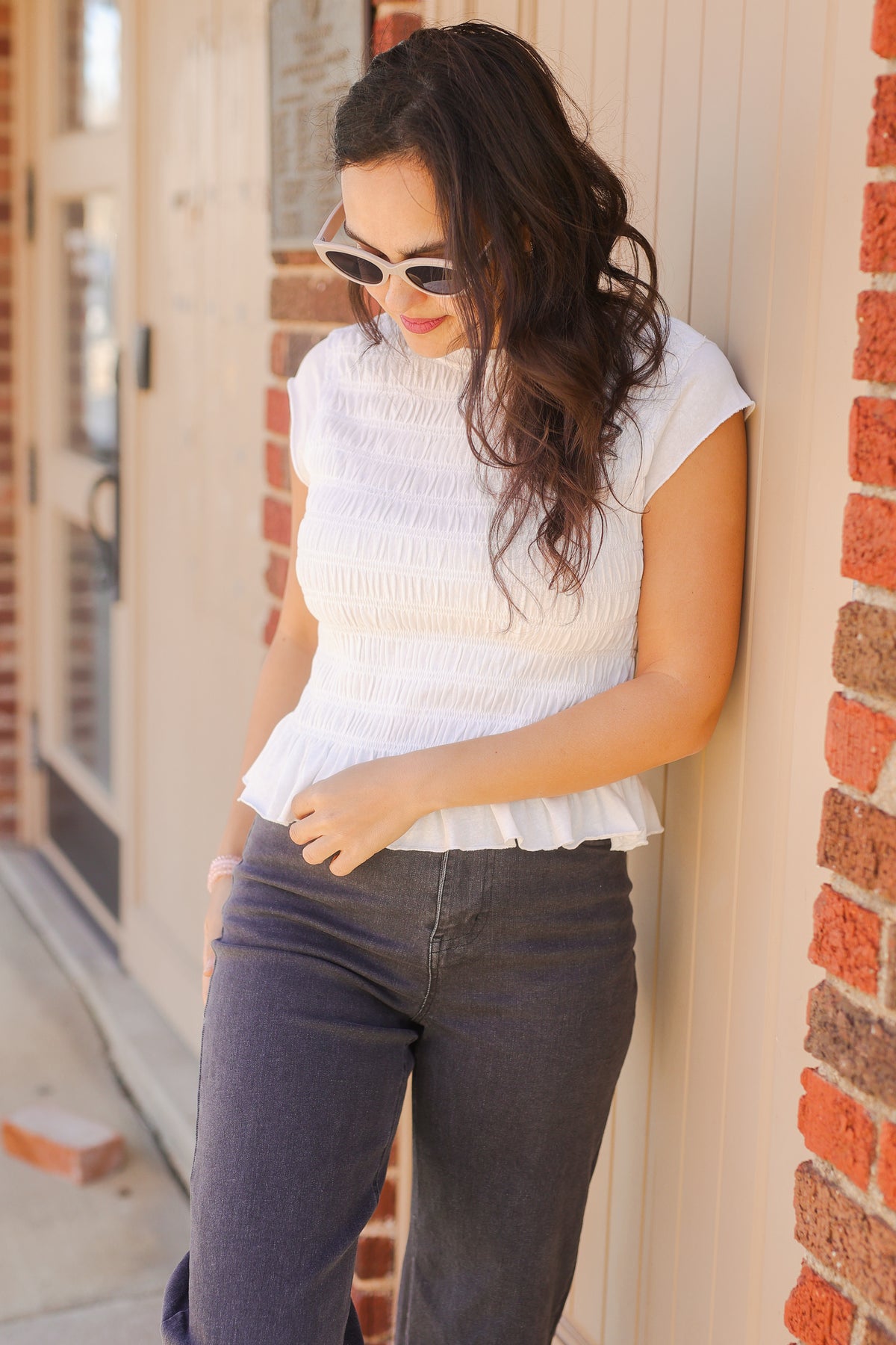 Woman wearing a white top and dark jeans leaning against a brick wall.