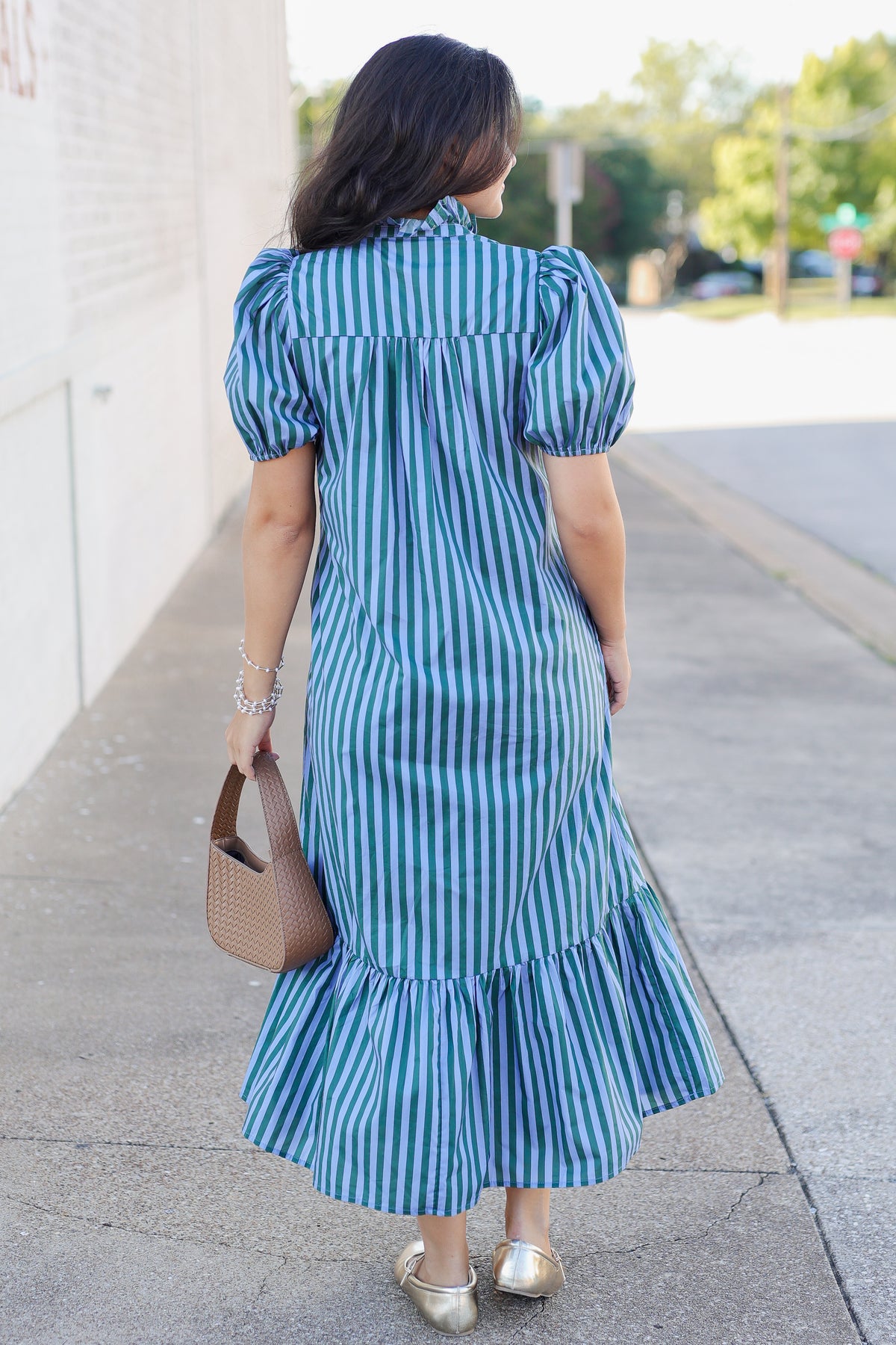 A woman viewed from behind wearing a blue and green vertically striped midi dress with puff sleeves and a ruffled hem, standing on a sidewalk next to a building.