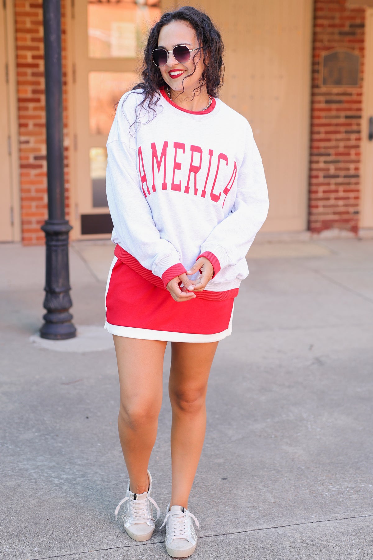 Woman wearing a white sweatshirt with 'AMERICA' in red and a red skirt on a sidewalk.