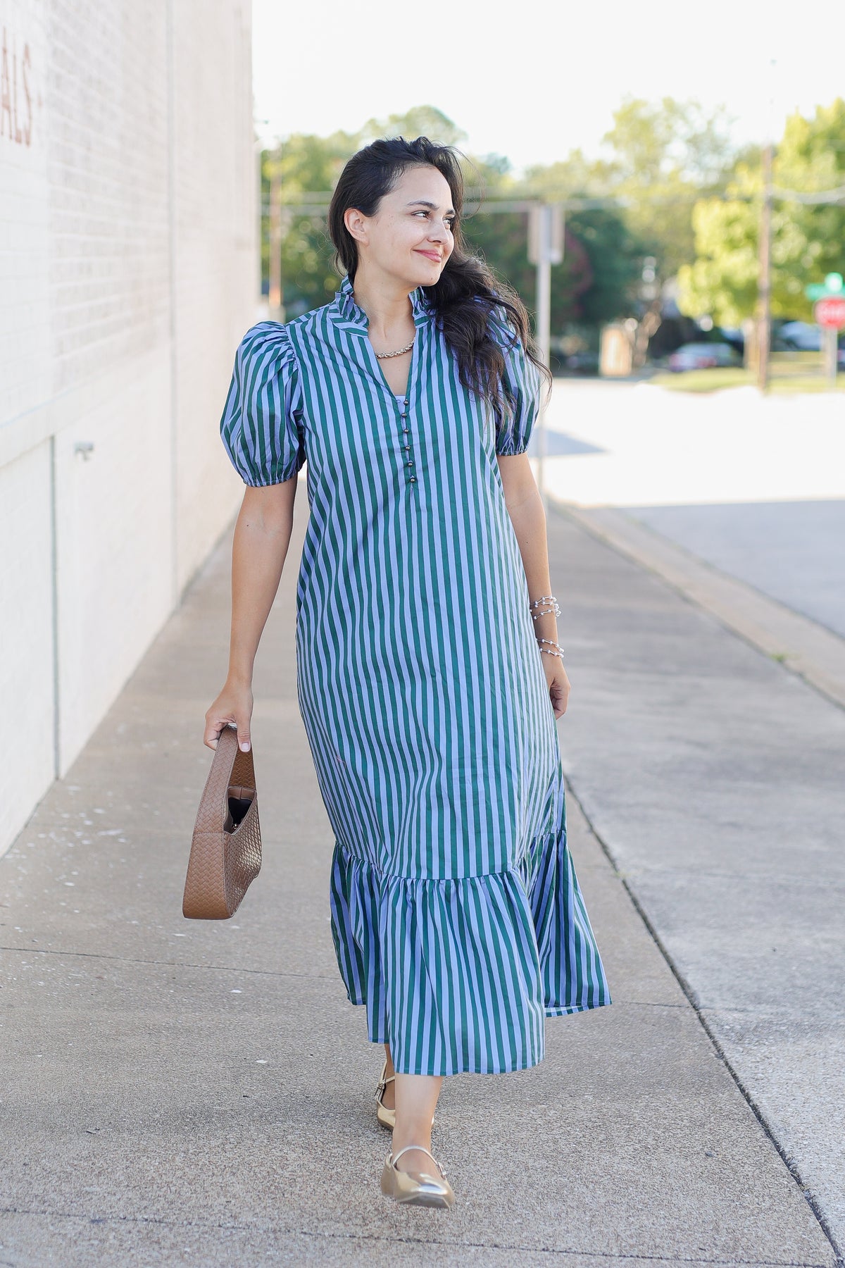 A woman wearing a blue and white vertically striped midi dress with puff sleeves walks on a sidewalk, holding a brown handbag.