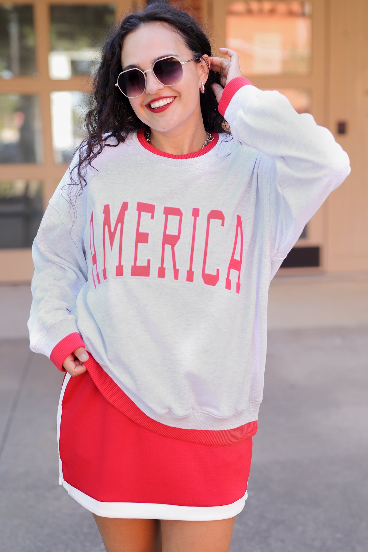 Woman wearing a white sweatshirt with 'AMERICA' printed in red and a red skirt outdoors.