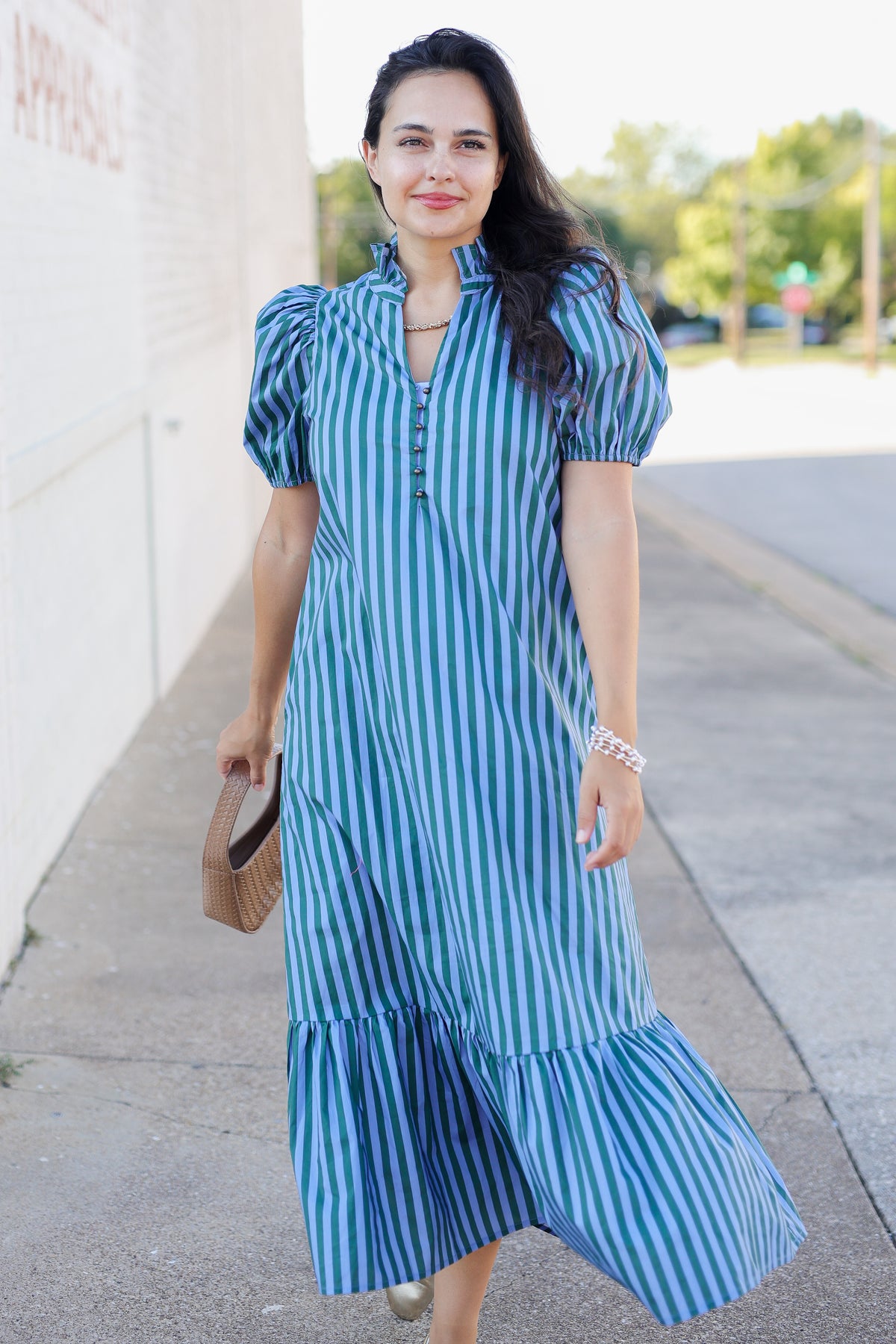 A woman wearing a blue and white vertically striped midi dress with puff sleeves and a ruffled hem, walking outdoors on a sidewalk next to a building.