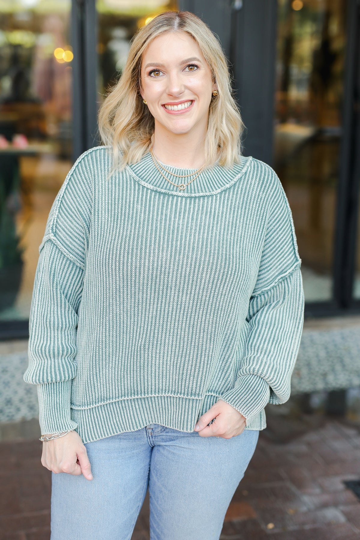 Woman wearing a green ribbed sweater and light blue jeans standing in front of a store.
