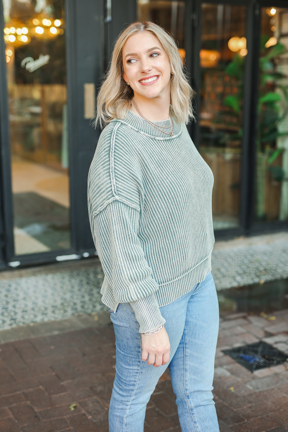 Woman wearing a green sweater and blue jeans standing on a sidewalk.