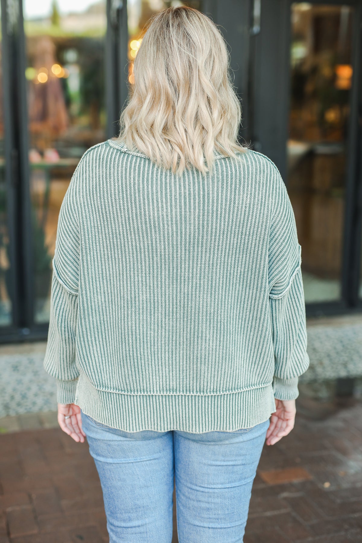 Person wearing a green ribbed sweater and blue jeans standing in front of a store entrance.