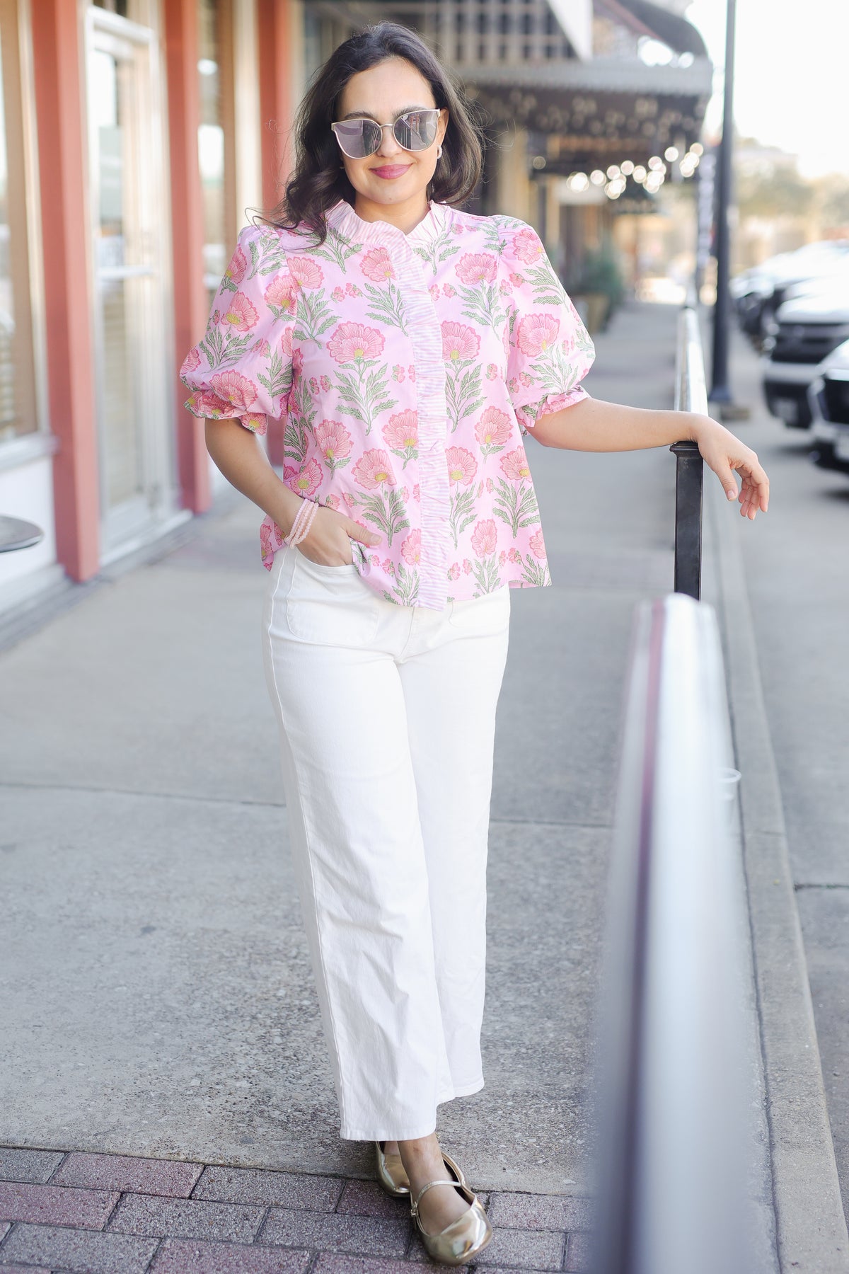 Woman wearing a pink floral blouse and white pants standing on a sidewalk.