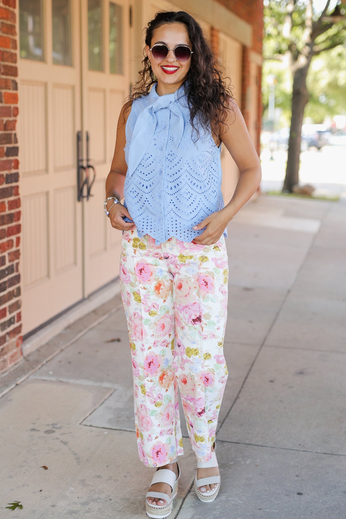 Woman wearing a light blue sleeveless top and floral pants on a sidewalk.