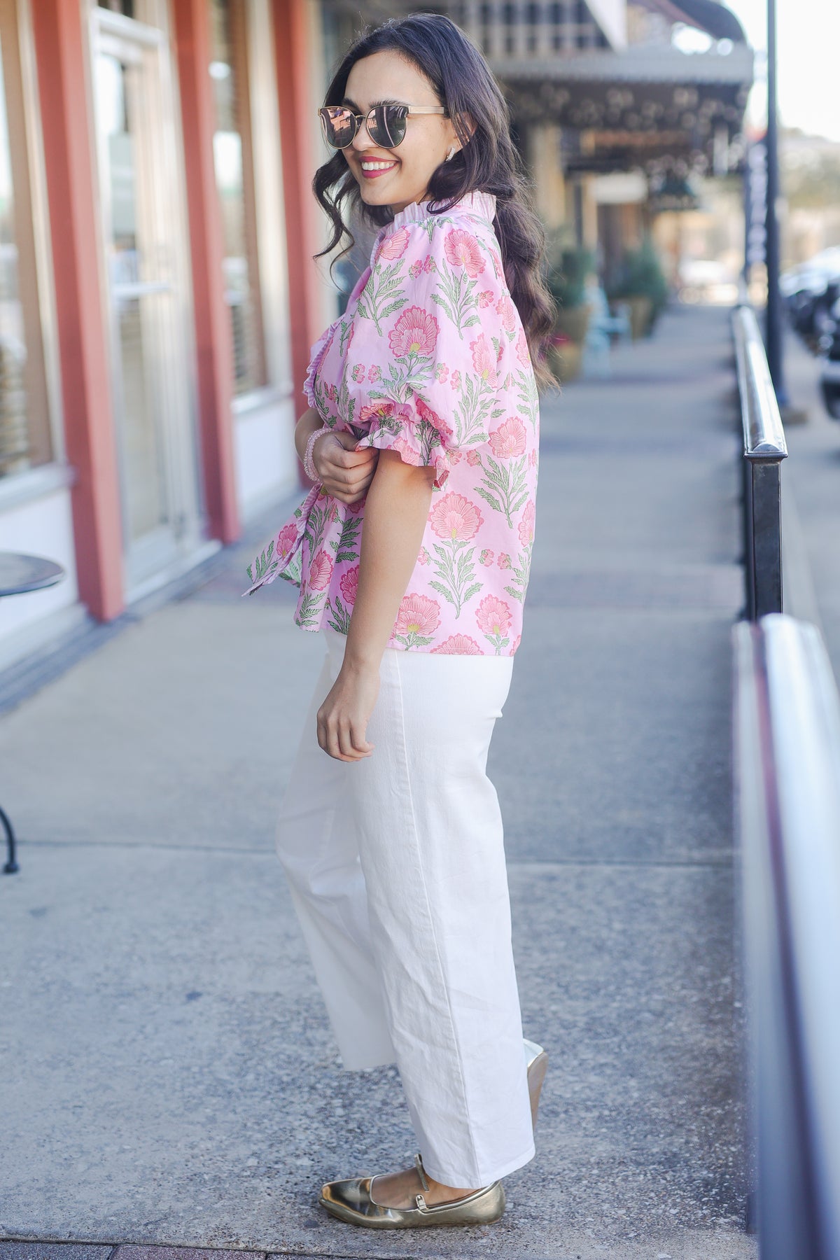 Woman wearing a floral blouse and white pants standing on a sidewalk.
