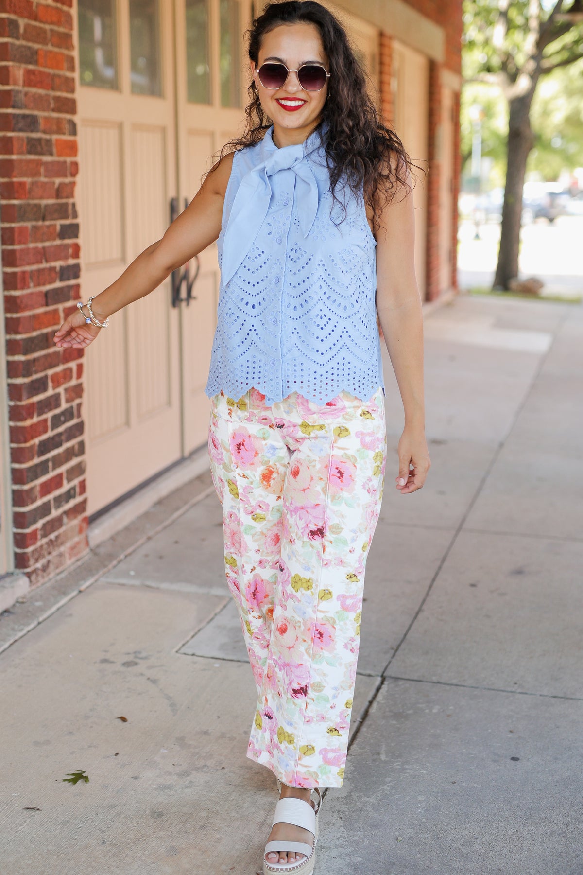 Woman wearing a light blue sleeveless top and floral pants on a sidewalk.