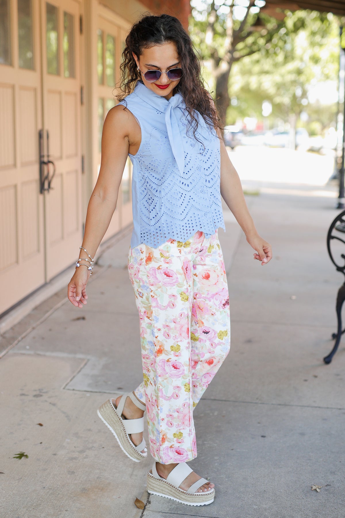 Woman wearing a light blue sleeveless top and floral pants on a sidewalk.