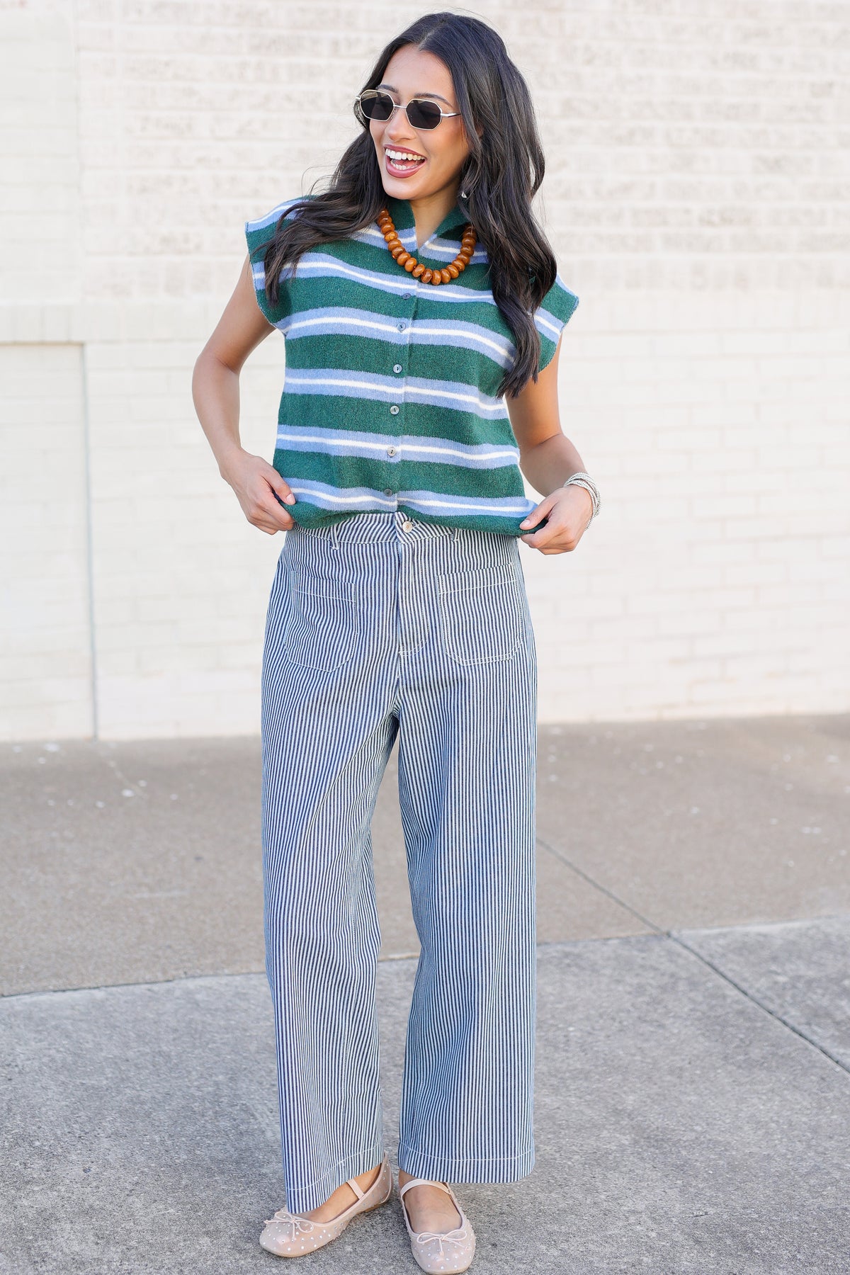 A woman wearing a green and white striped short-sleeve button-up top, light blue pinstriped wide-leg pants, and beige flats, standing outdoors against a light brick wall.