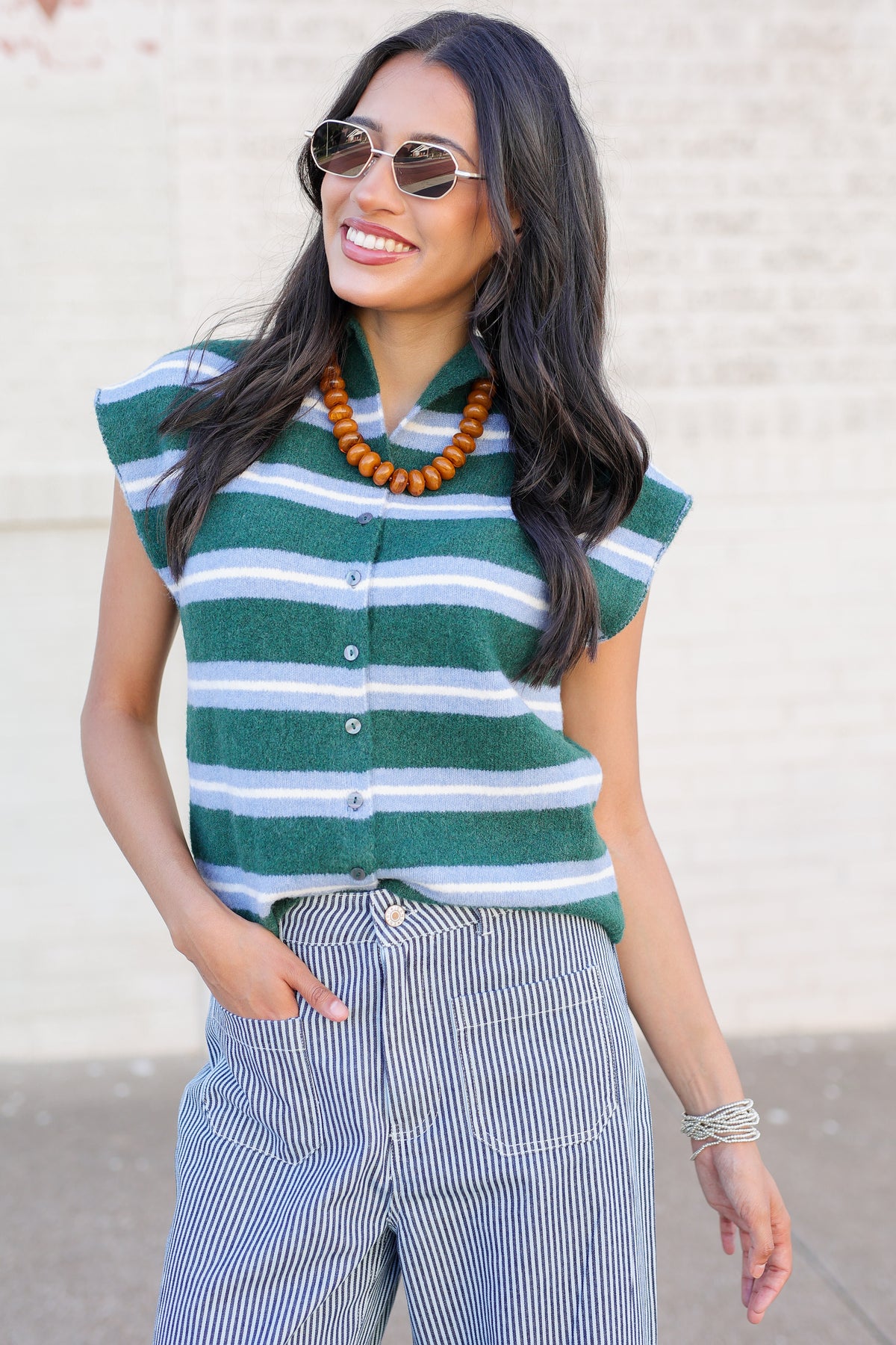 A woman wearing a green and white striped button-down knit top, blue and white pinstriped pants, sunglasses, and a chunky wooden bead necklace, standing against a light brick wall.