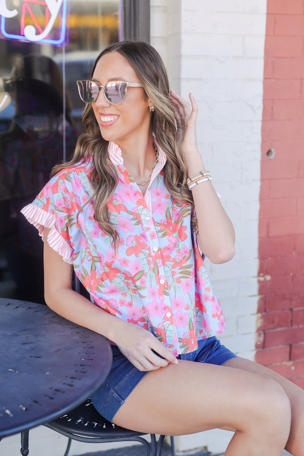 Woman wearing a colorful floral blouse and denim shorts sitting outdoors.