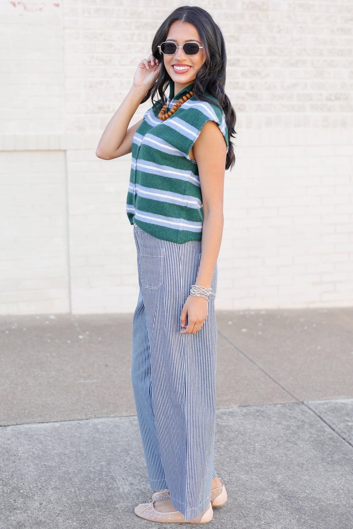 A woman modeling a green and white striped knit top with light blue pinstriped wide-leg pants, styled with sunglasses, a beaded necklace, and flat shoes against a light brick wall.